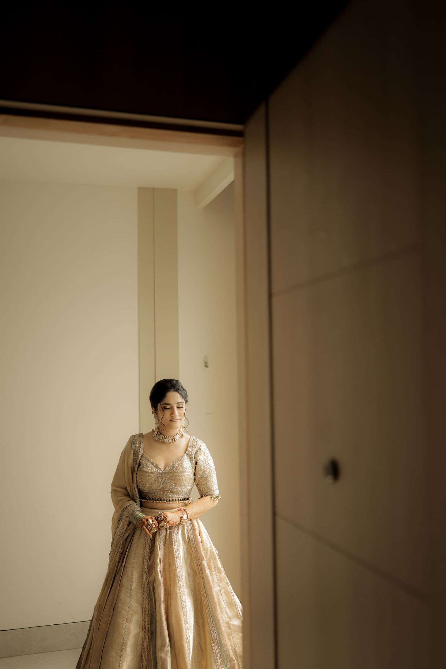 A bride in a golden lehenga stands elegantly in a warmly lit doorway, exuding grace and poise.