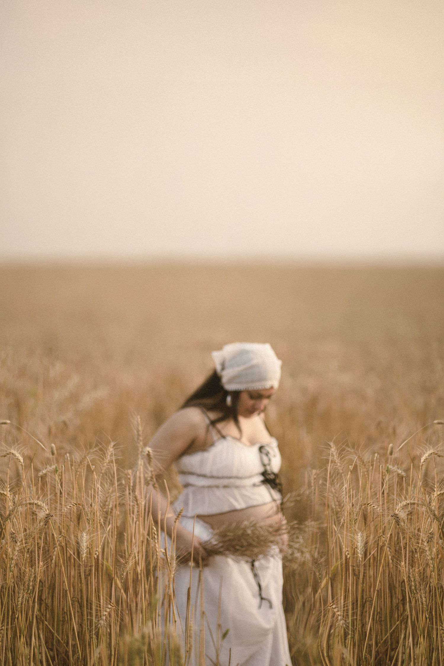Pregnant woman in white dress and headscarf standing in golden wheat field, cradling her baby bump.