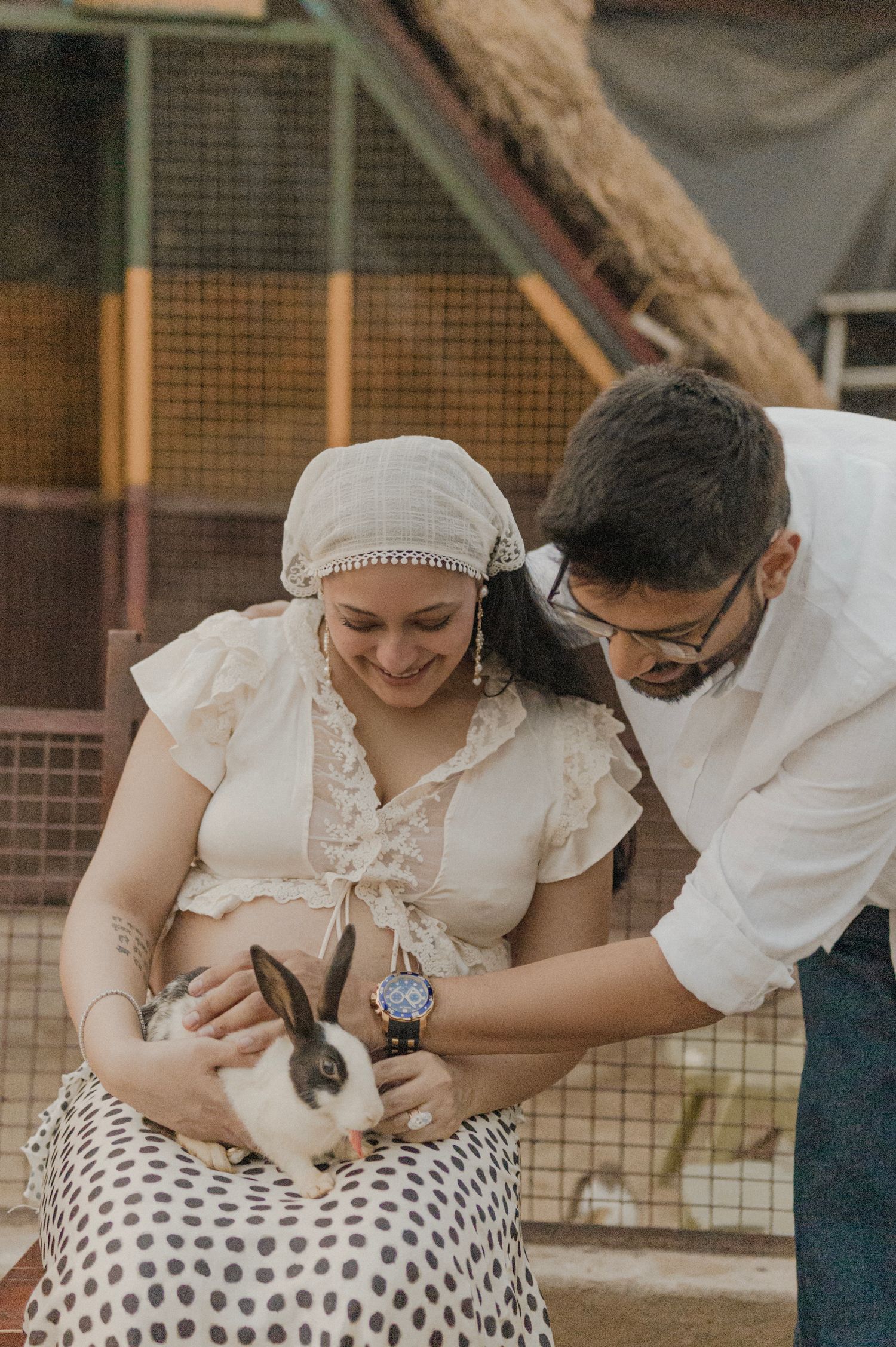 Pregnant woman in white outfit holds a bunny while a man in white shirt leans in, both smiling warmly.