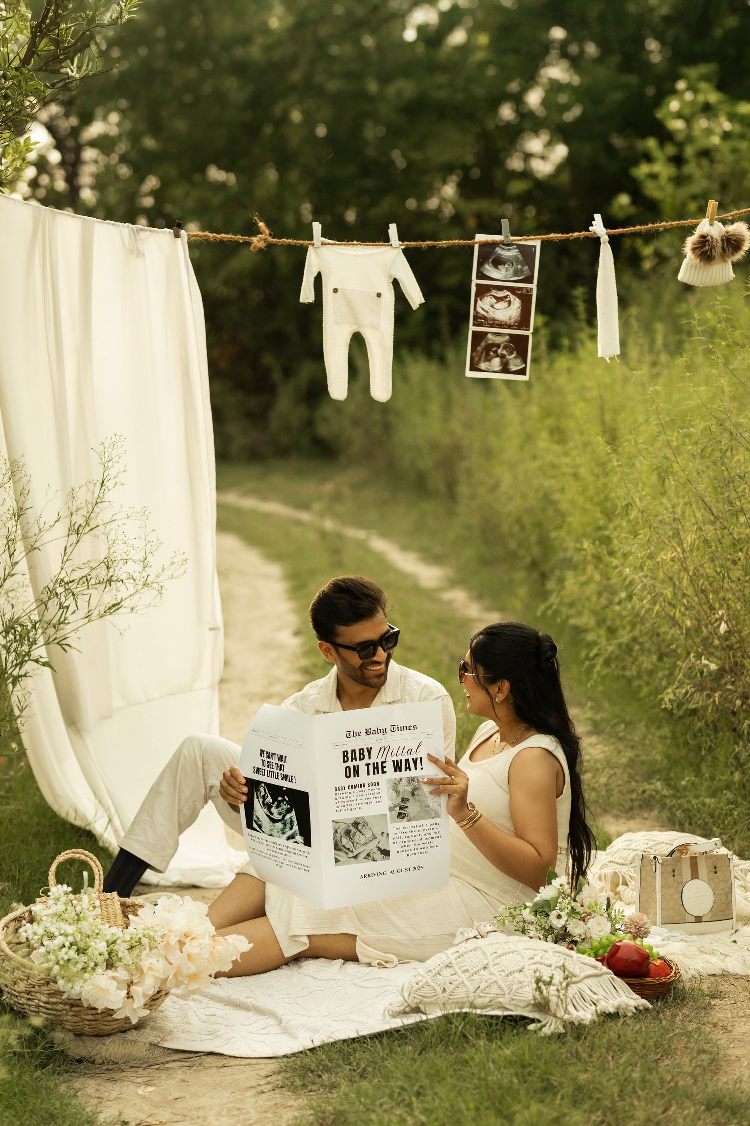 Couple sitting on picnic blanket outdoors with baby announcement newspaper, ultrasound photos, and tiny onesie hanging on clothesline.