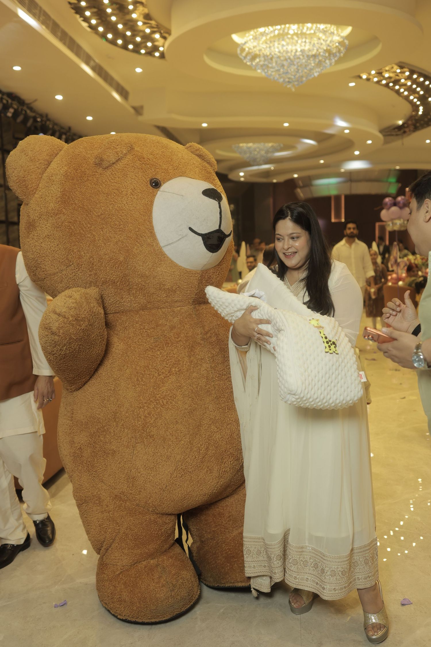 Woman in a cream embroidered outfit holding a swaddled baby beside a giant teddy bear costume in an elegant banquet hall with chandeliers.