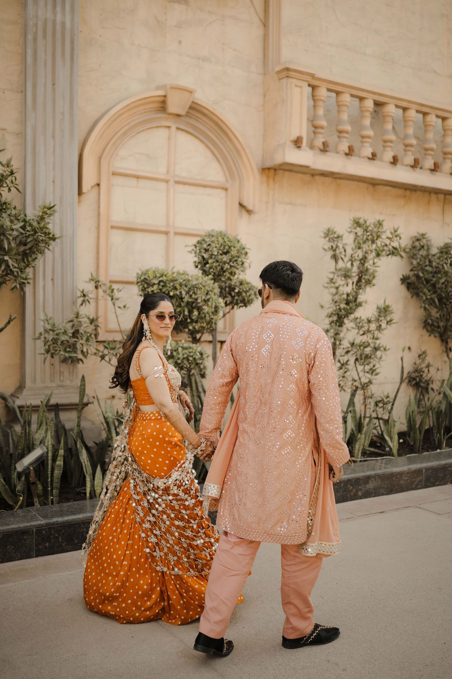 Couple in traditional Indian attire, woman in orange lehenga and man in pink sherwani, holding hands outside ornate building.