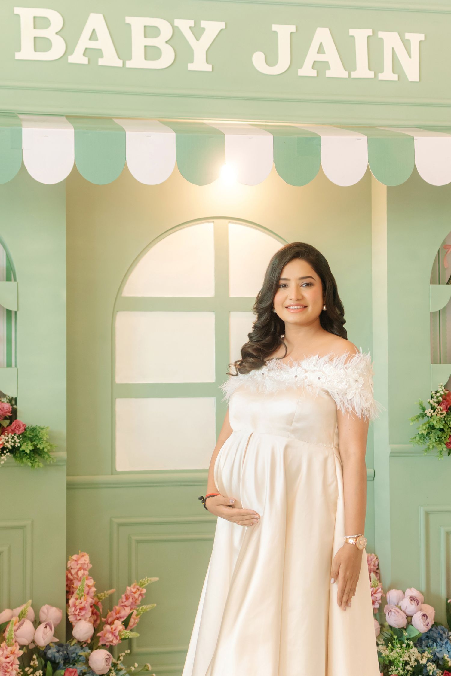 Pregnant woman in white off-shoulder gown poses at Baby Jain baby shower backdrop with floral decorations.