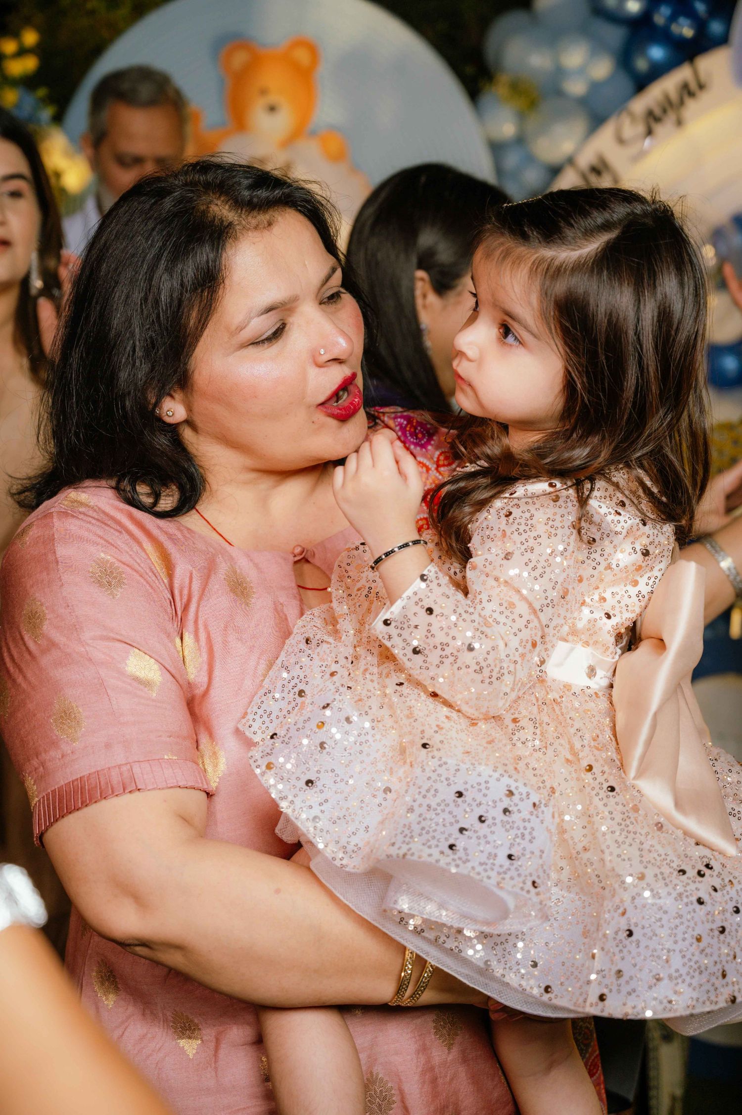 Woman in pink dress holding a toddler girl in a sparkly white dress at a birthday party celebration.