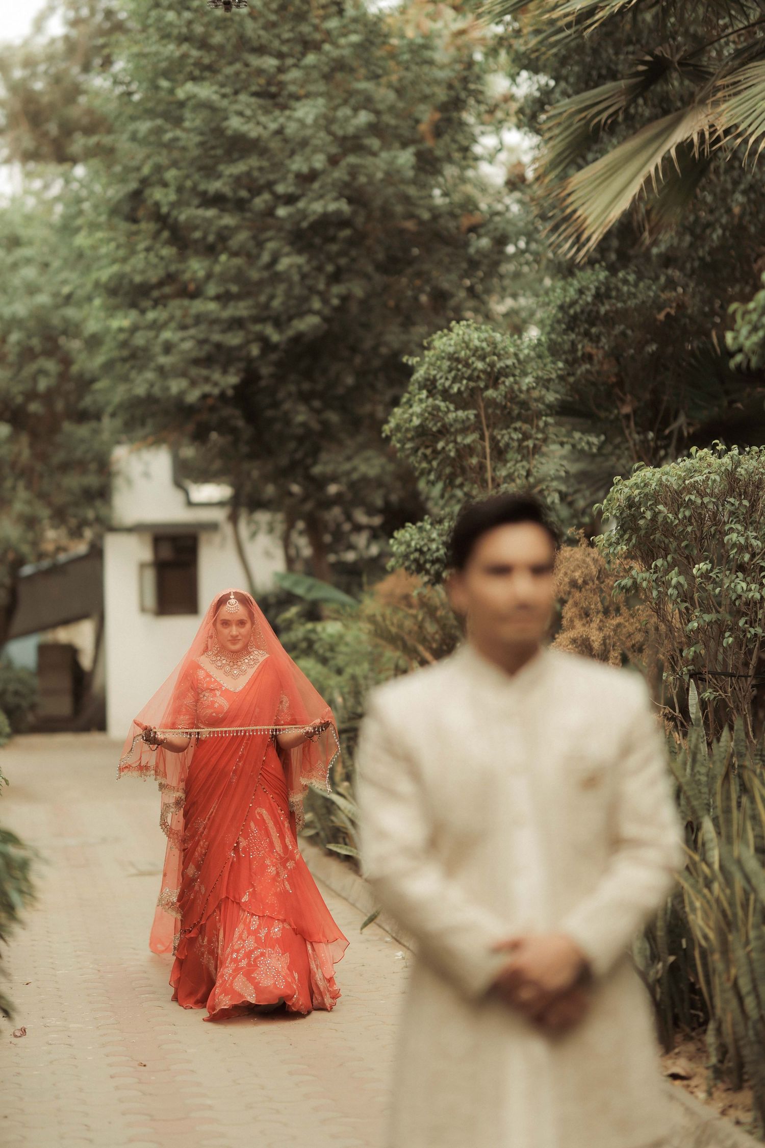 A bride in a red lehenga with veil walks toward a groom in white sherwani on a garden path.