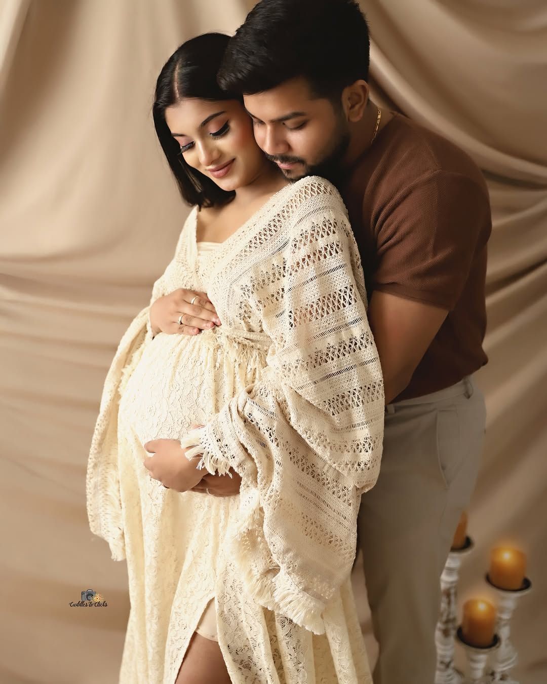 Intimate maternity photo with couple in cream and brown tones against draped backdrop.