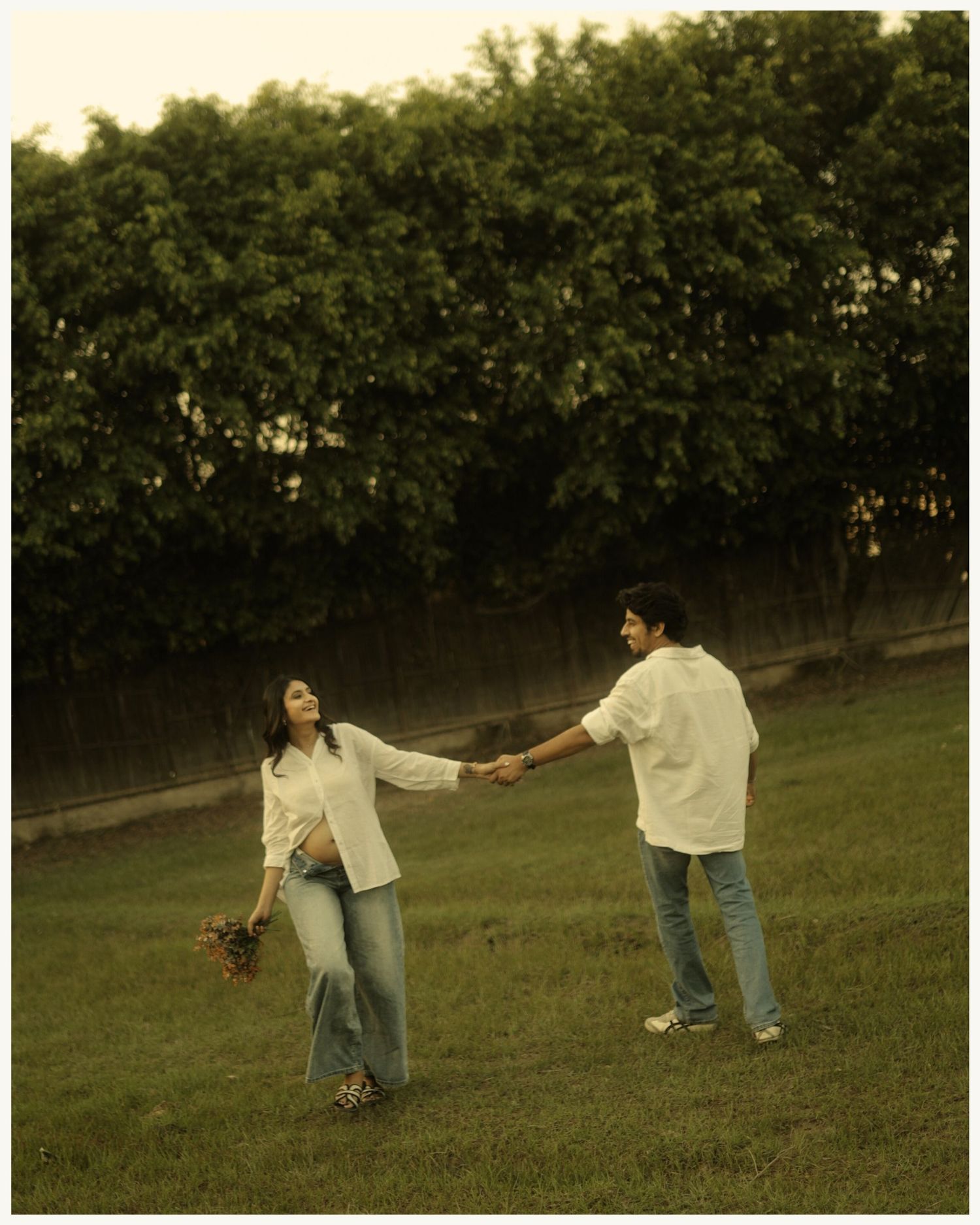 Couple holding hands and dancing playfully in a green meadow, wearing casual white shirts and jeans.