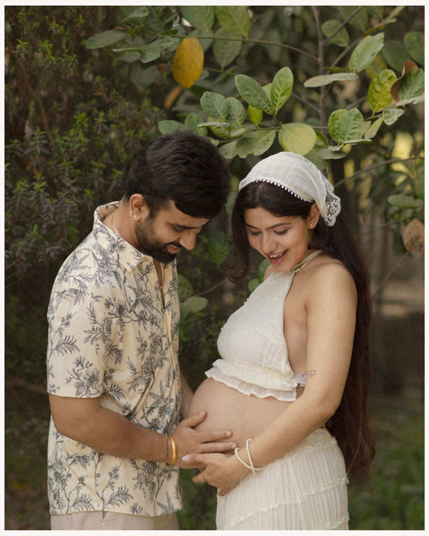 A couple tenderly cradles a pregnant belly outdoors, surrounded by lush green foliage in a maternity photoshoot.