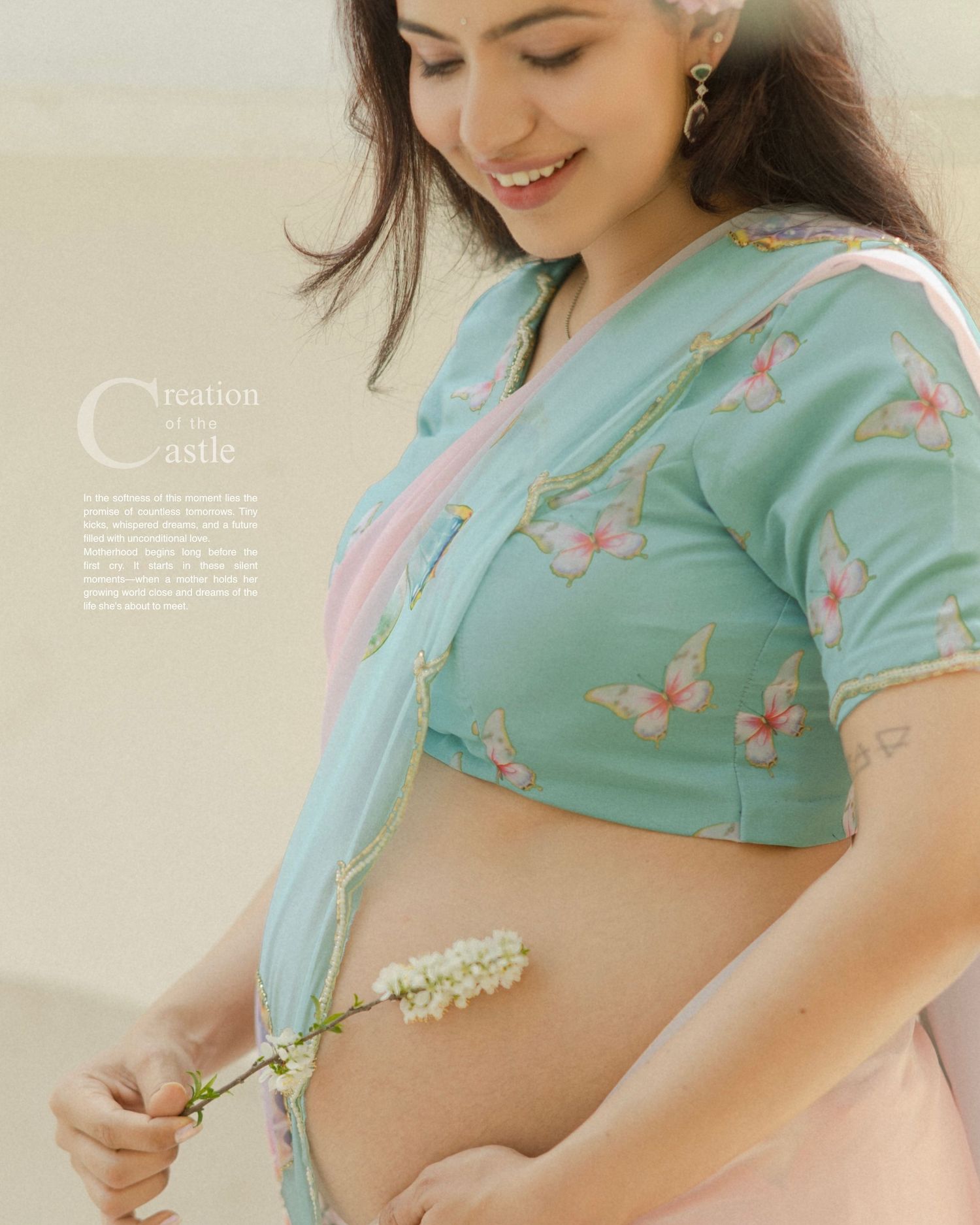 Pregnant woman in teal butterfly blouse and pink saree holding white flowers, smiling during maternity photoshoot.