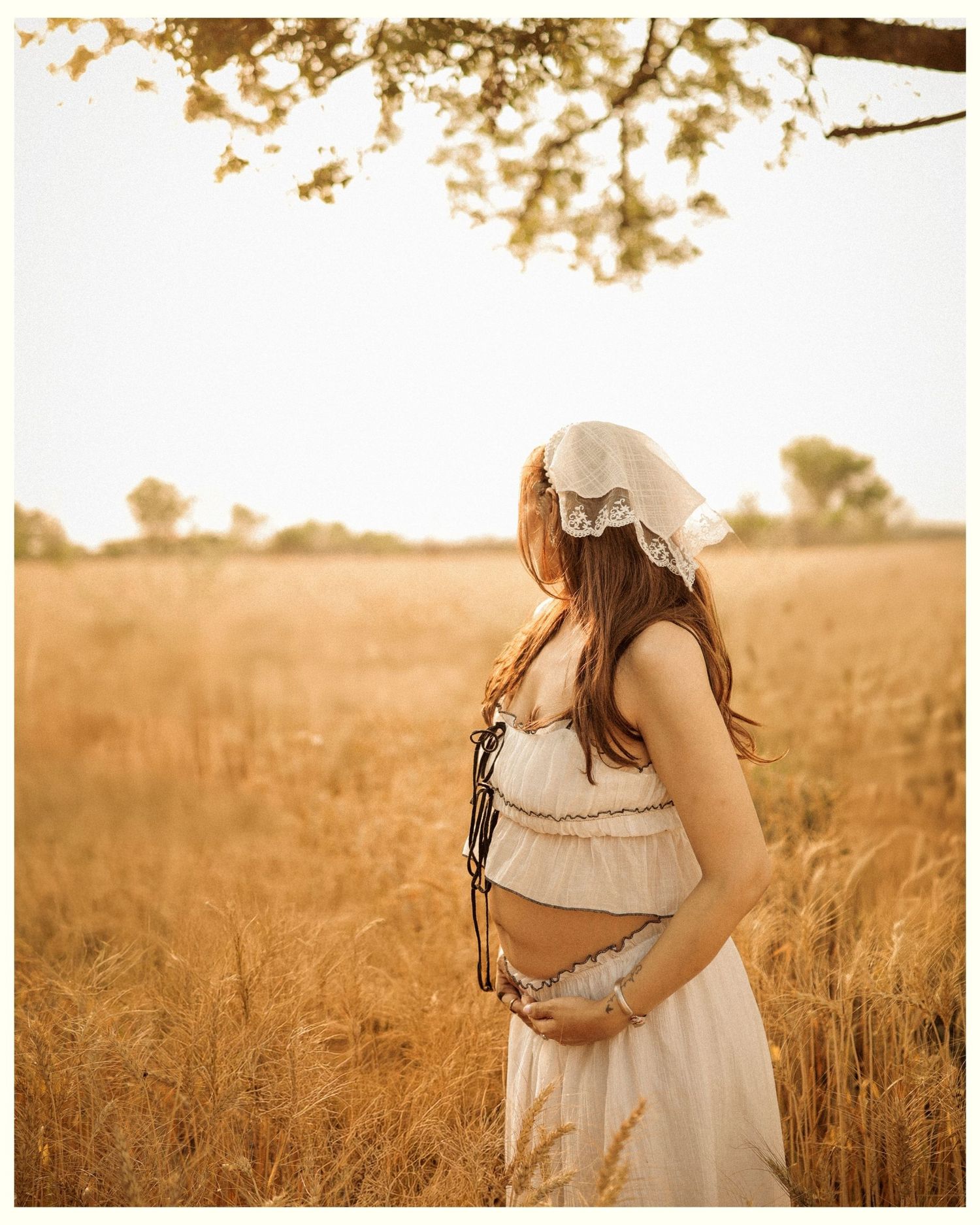 Pregnant woman in white dress and lace bonnet standing in golden wheat field during sunset.