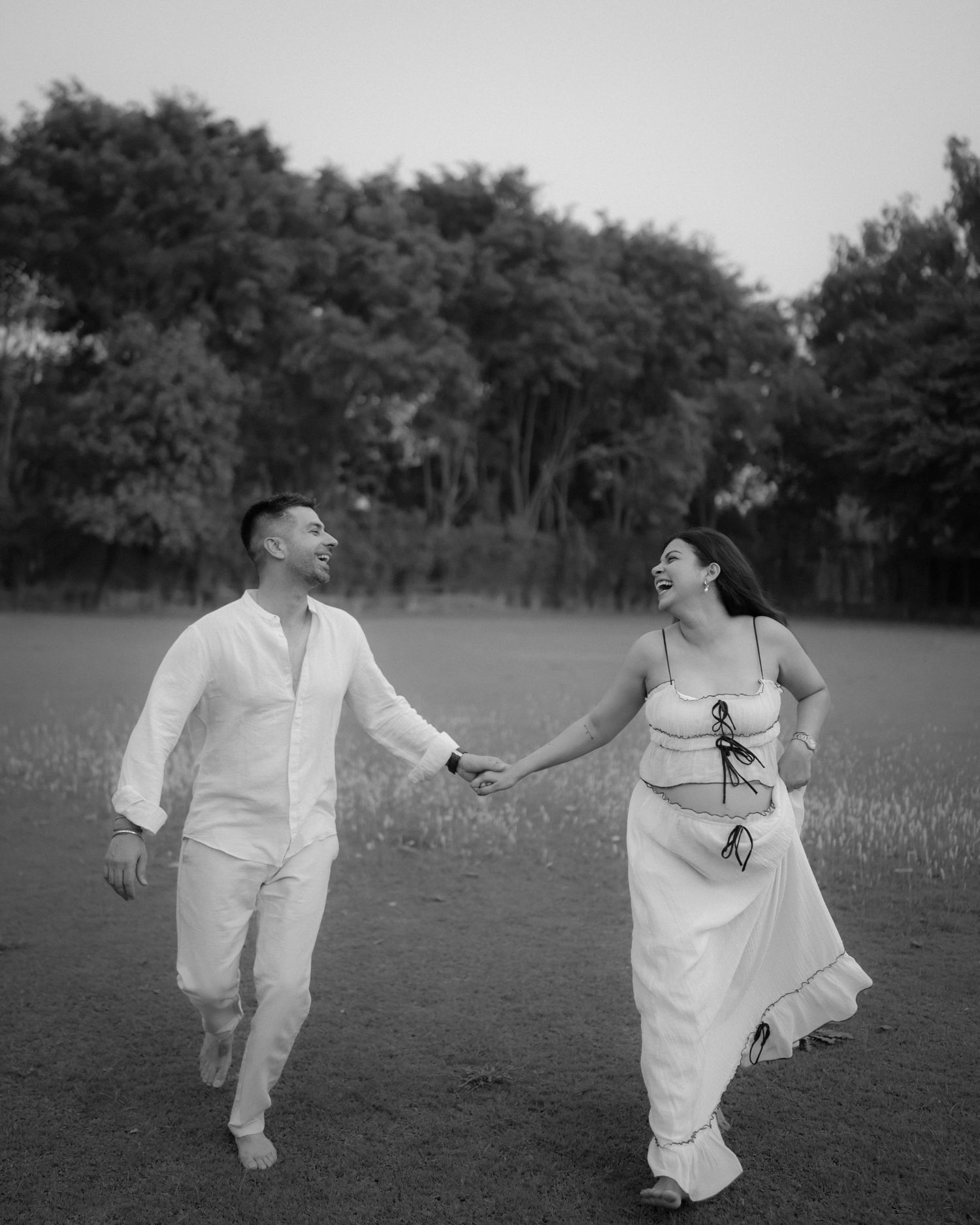 Black and white photo of a couple in white outfits holding hands and laughing while walking in a field.
