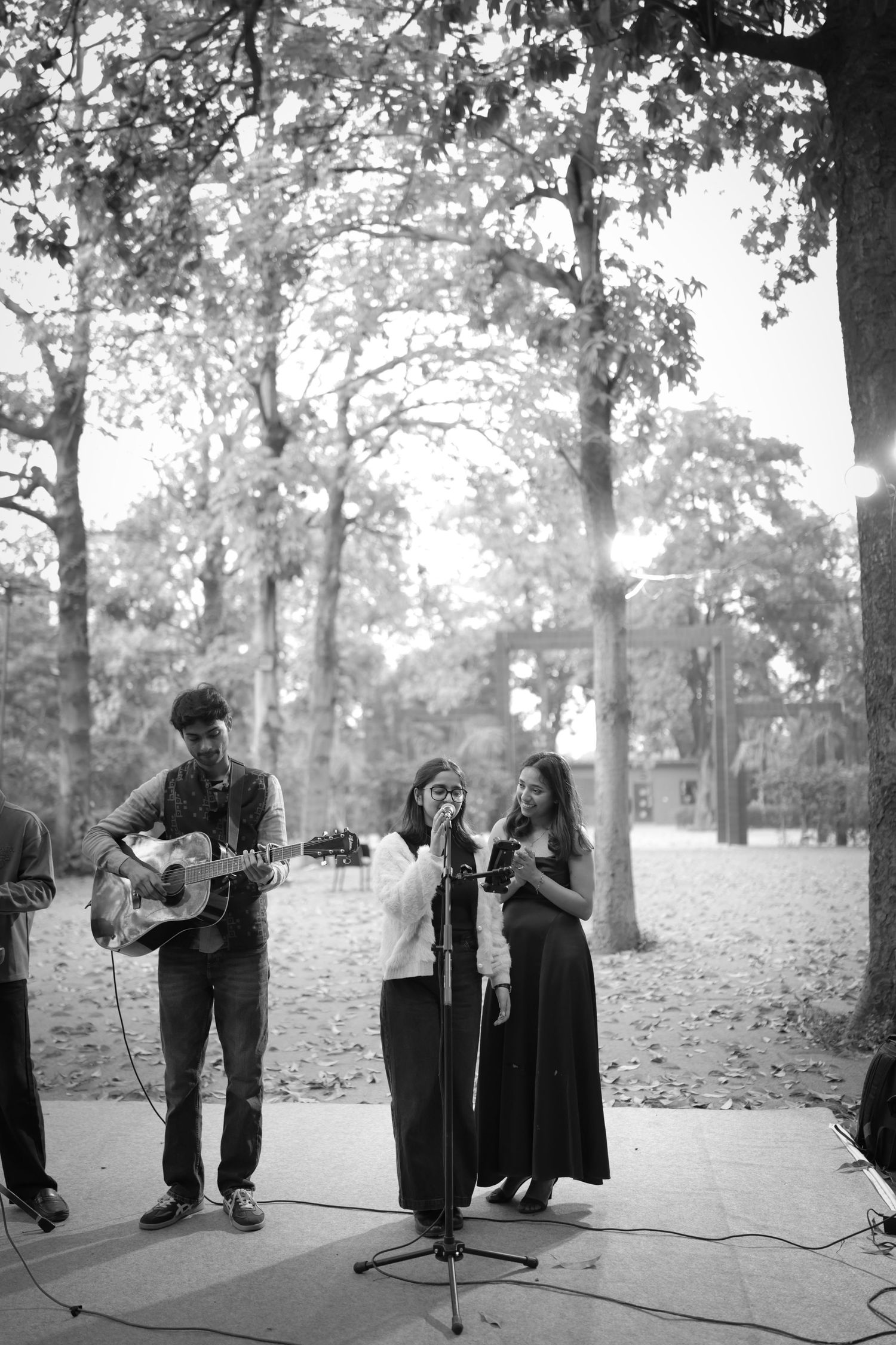 Black and white photo of three musicians performing outdoors under trees, with a guitarist and two singers at a microphone.