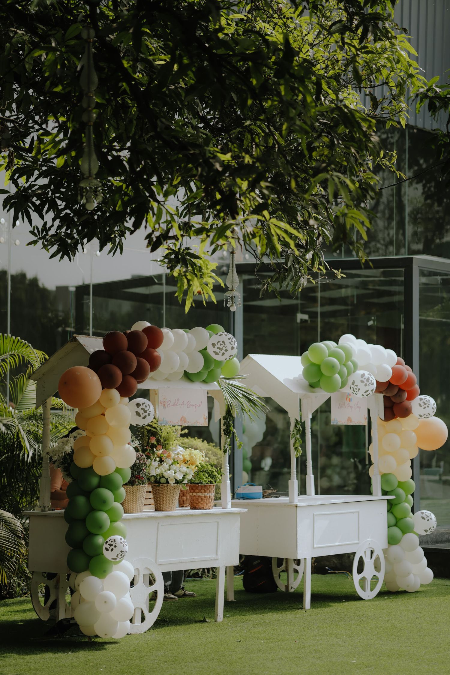 White cart decorated with colorful balloons in green, white, and burgundy, set on a lawn with lush trees in the background.