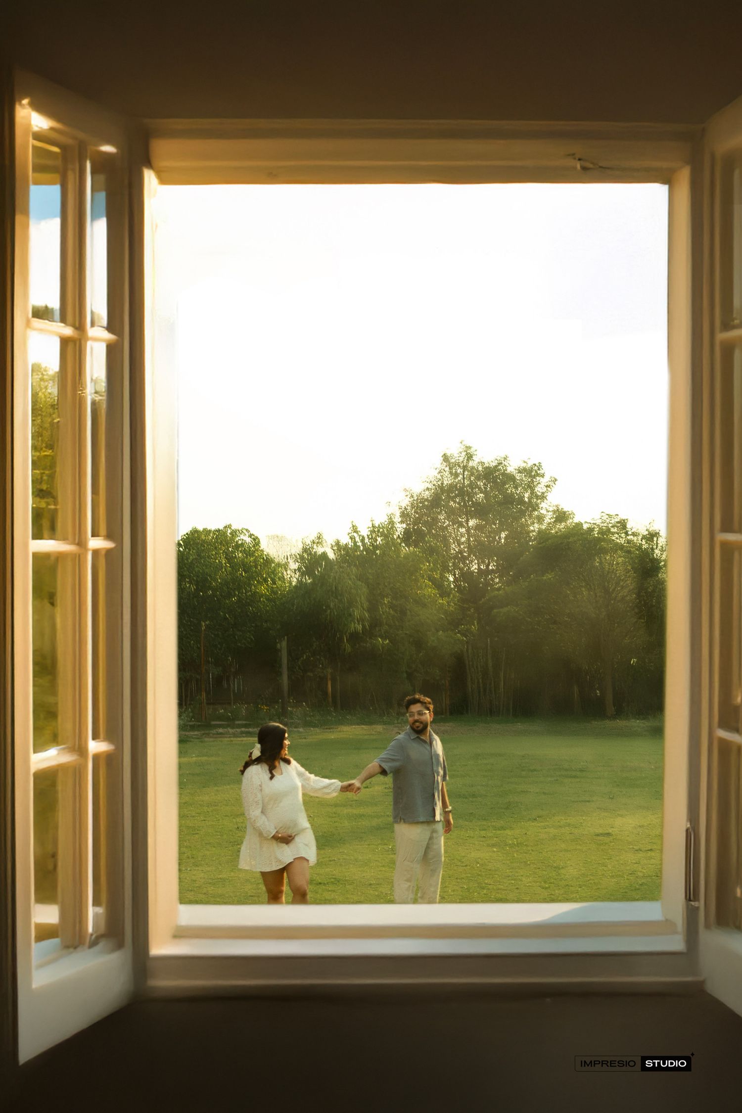 Couple holding hands in golden hour sunlight viewed through open white French doors with lush green lawn backdrop.