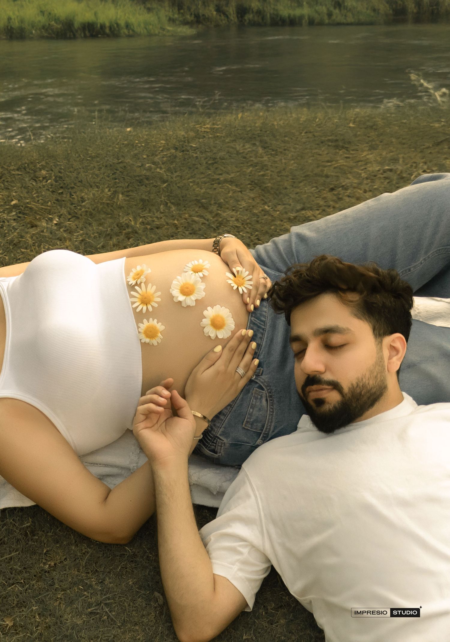Couple lying outdoors by a river, man resting head near pregnant woman's belly decorated with daisy flowers.