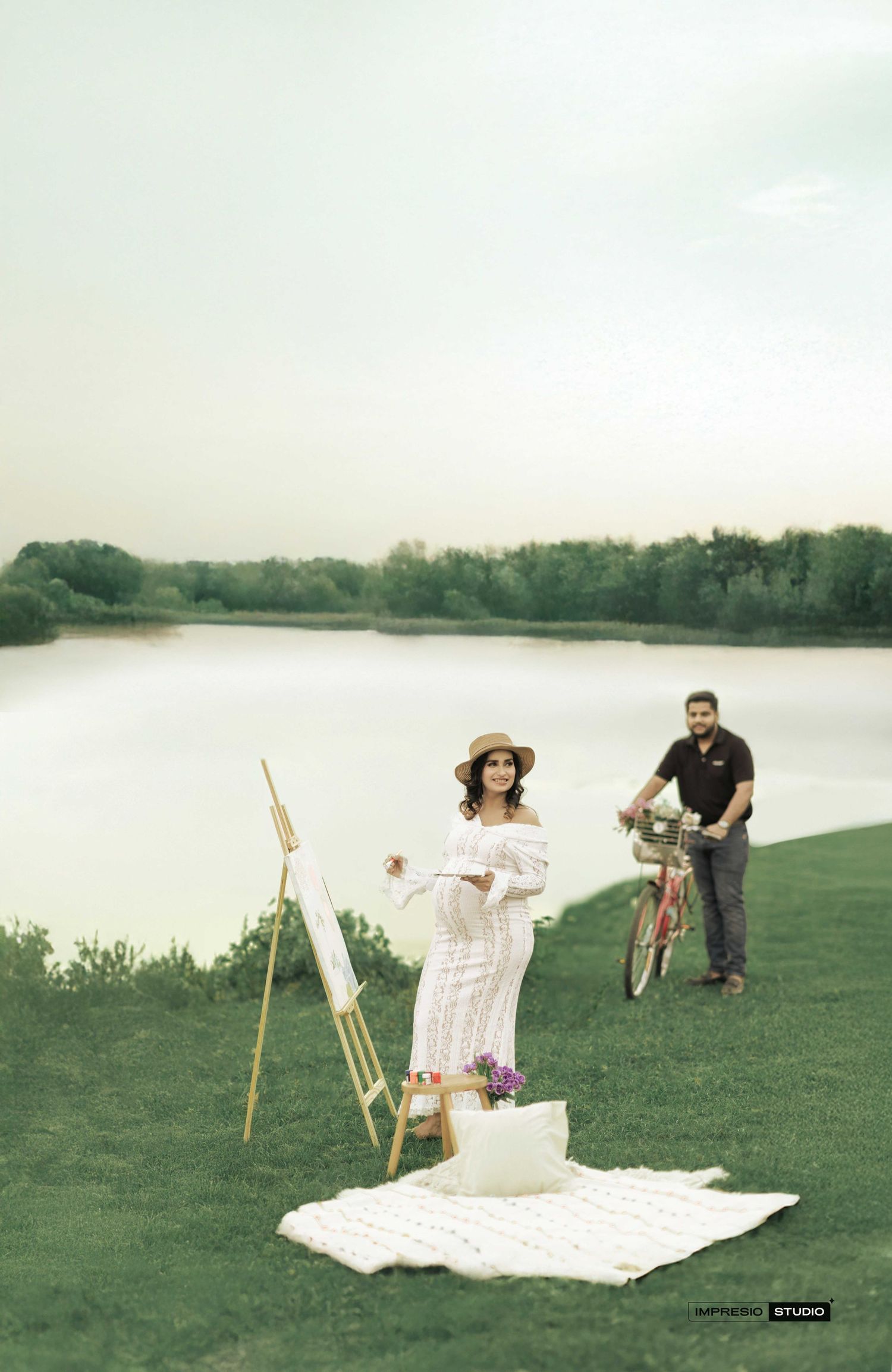 Pregnant woman in white dress painting by easel near lake, man with bicycle in background during maternity photoshoot.