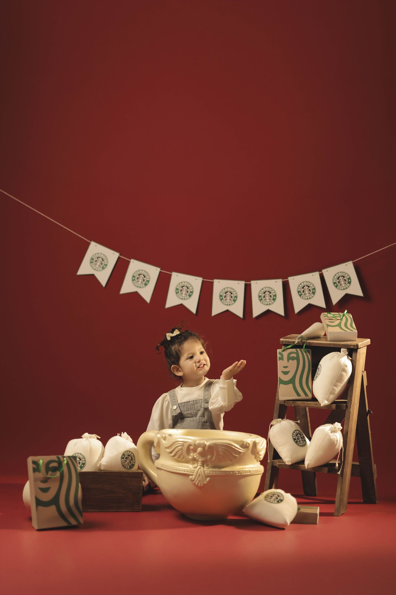 A toddler girl sits beside a giant Starbucks cup with branded decor on a festive red background.