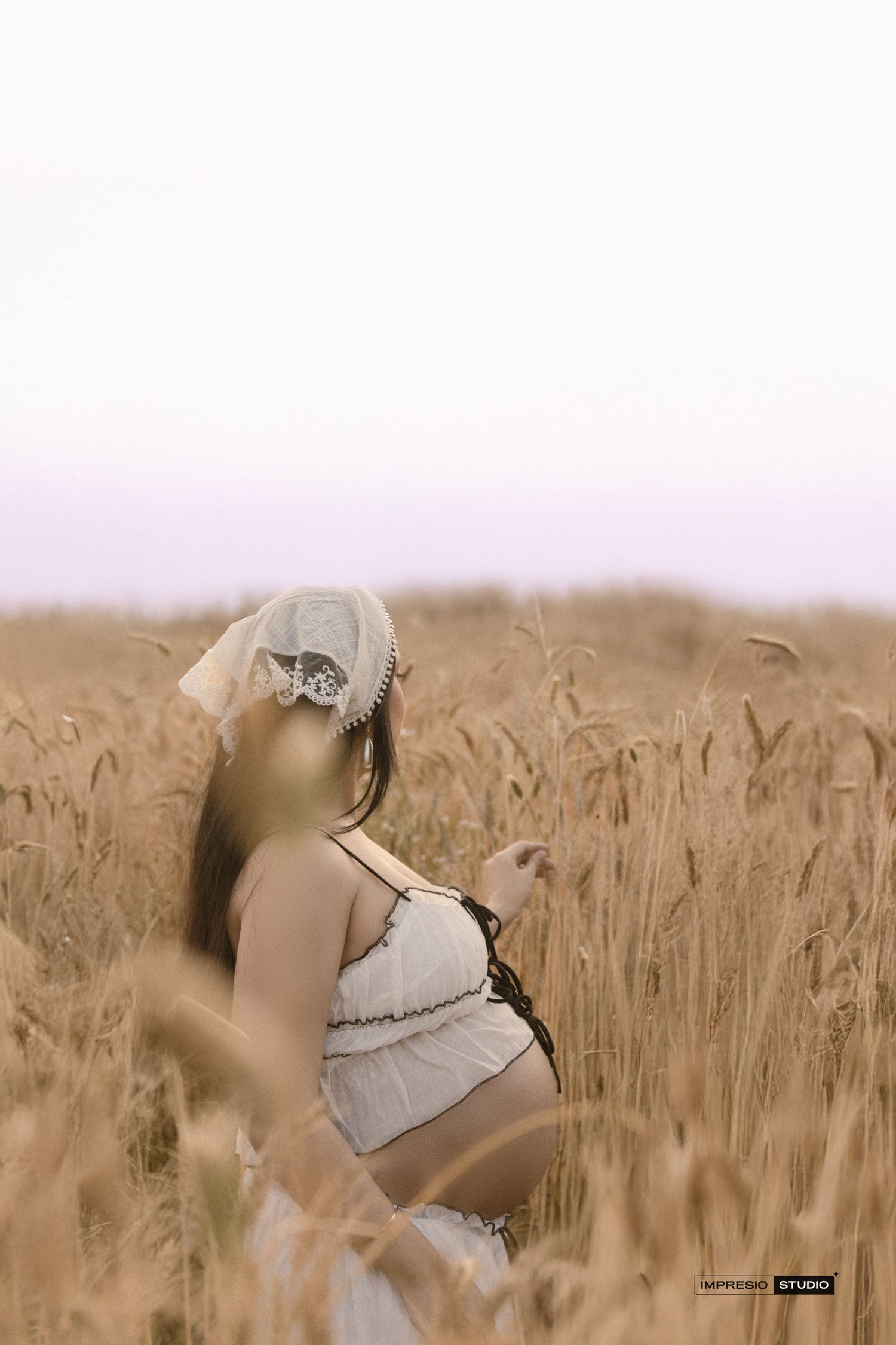 Pregnant woman in white dress and hat standing in golden wheat field during maternity photoshoot.