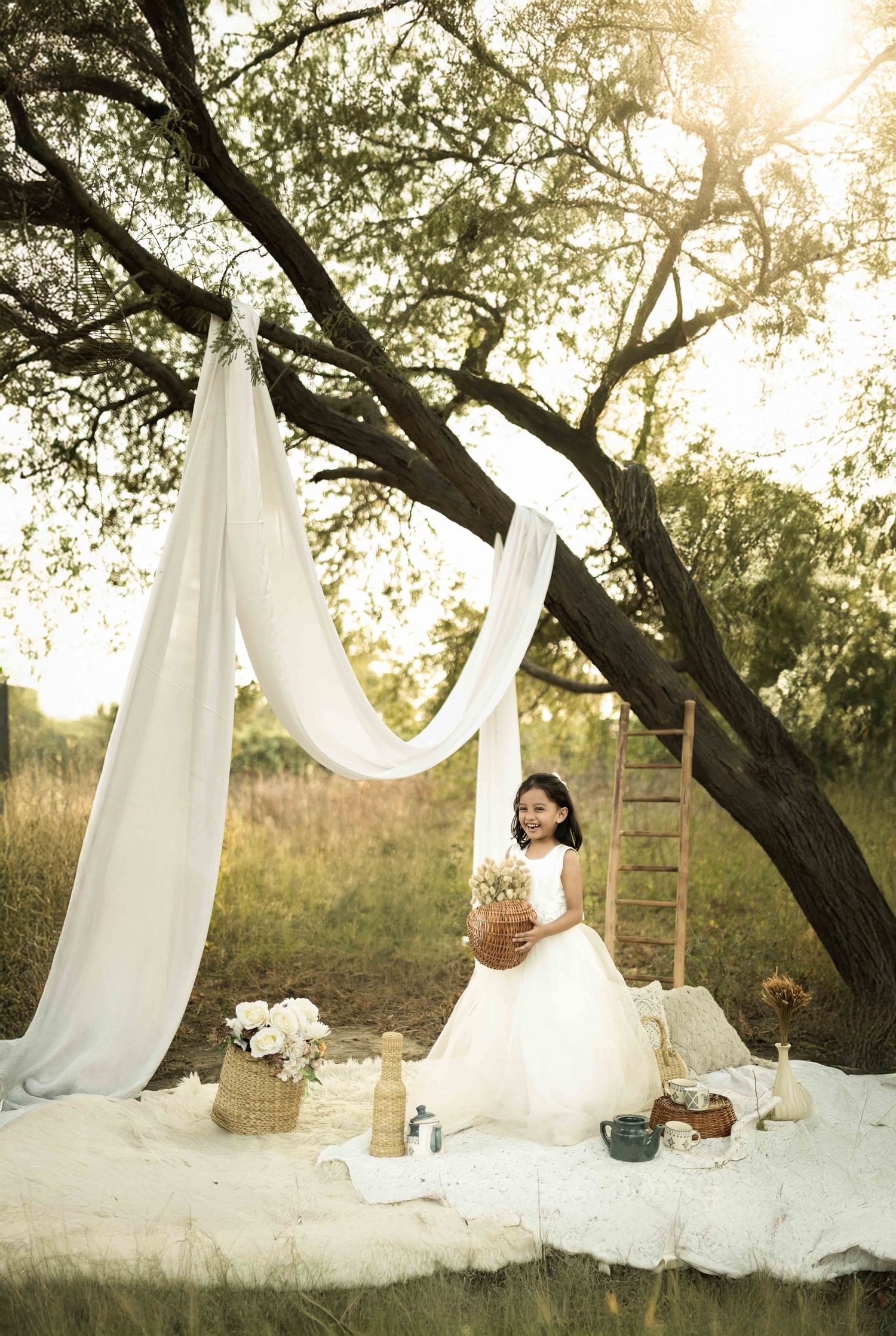Young girl in white dress poses under draped fabric canopy outdoors during golden hour for a whimsical portrait session.