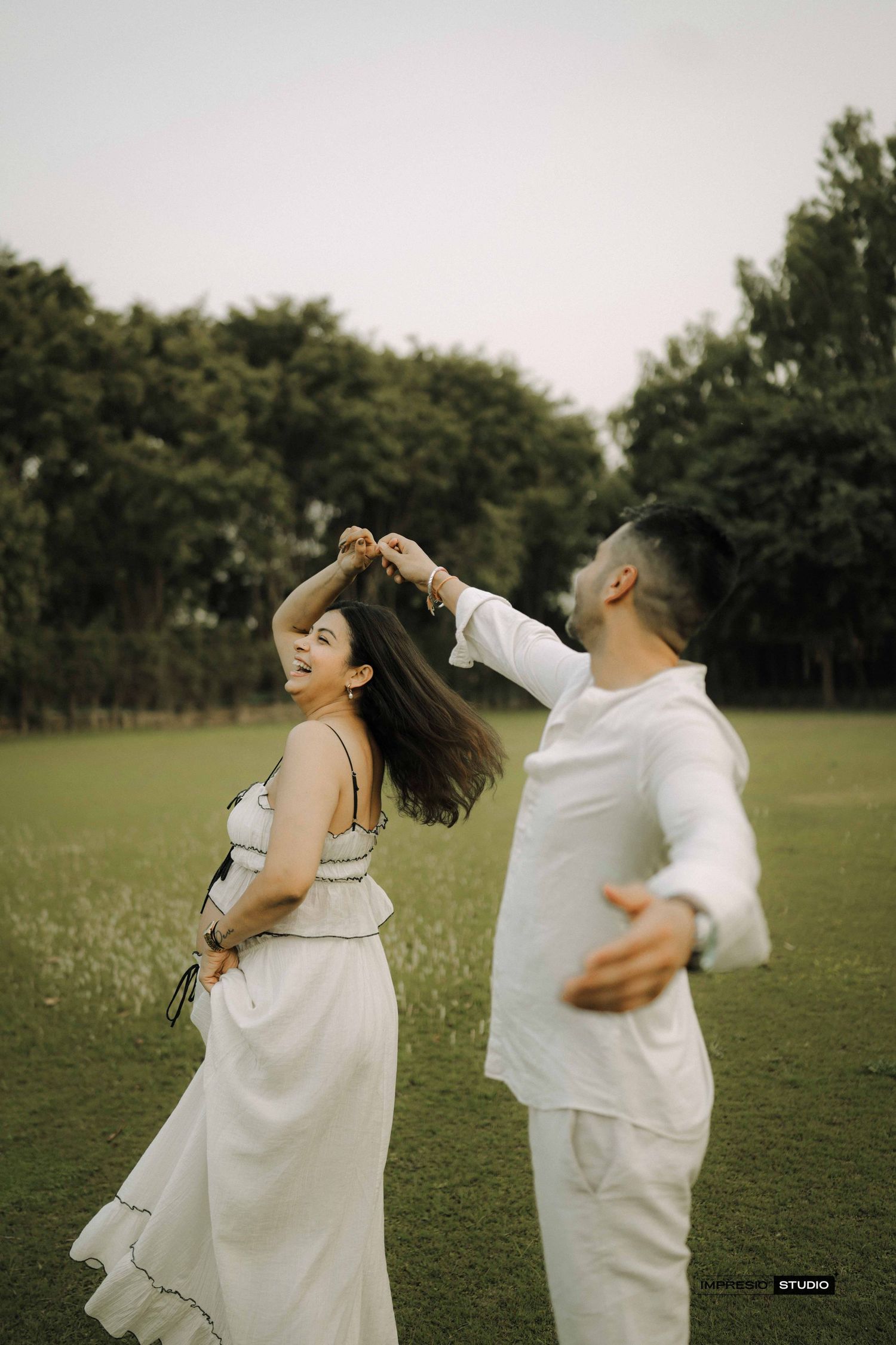 Couple dancing in a green field, woman in white dress spinning joyfully as man in white outfit twirls her outdoors.