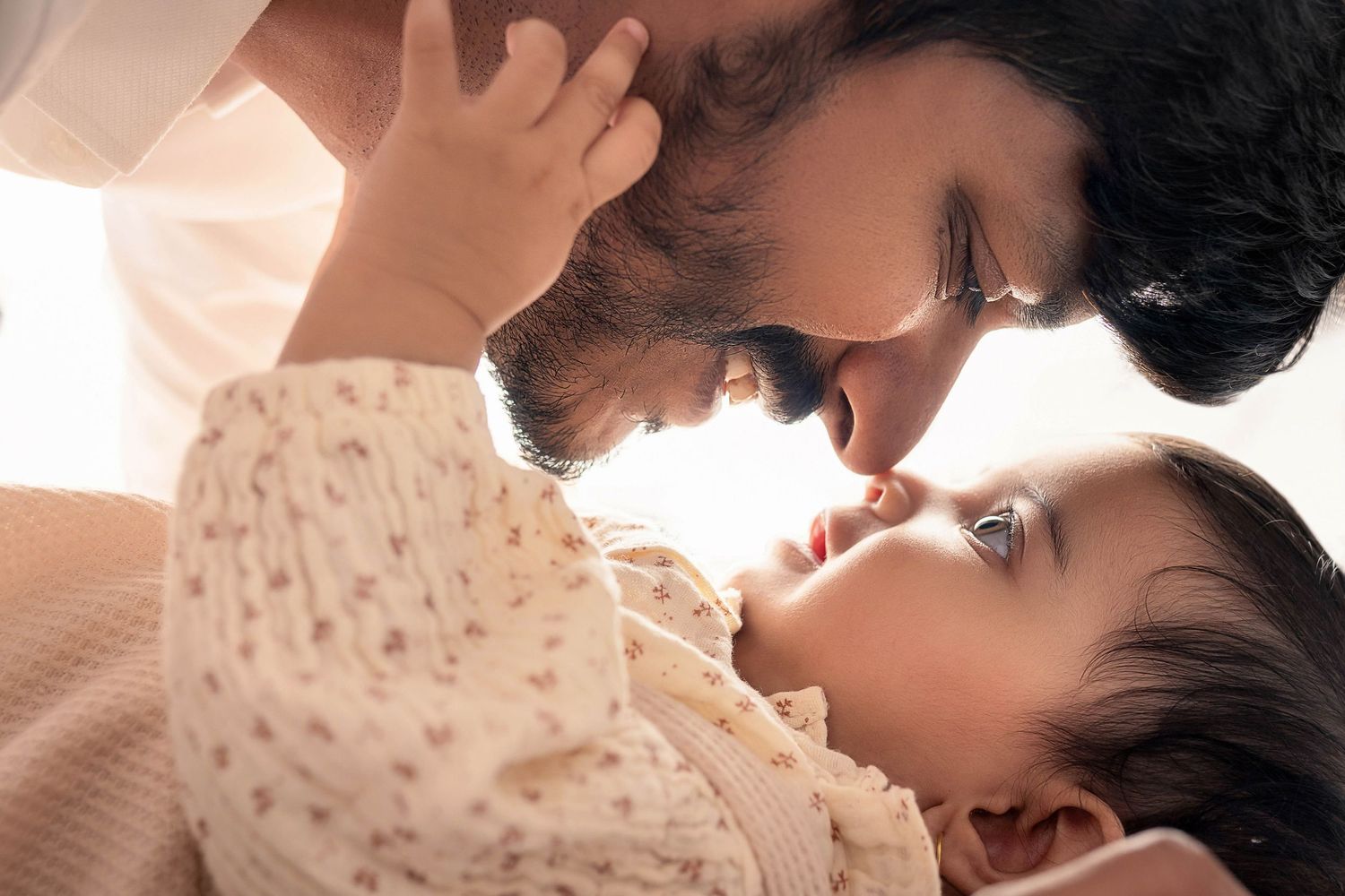 A tender moment between a bearded father and his newborn baby, sharing a gentle nose-to-nose touch in soft light.