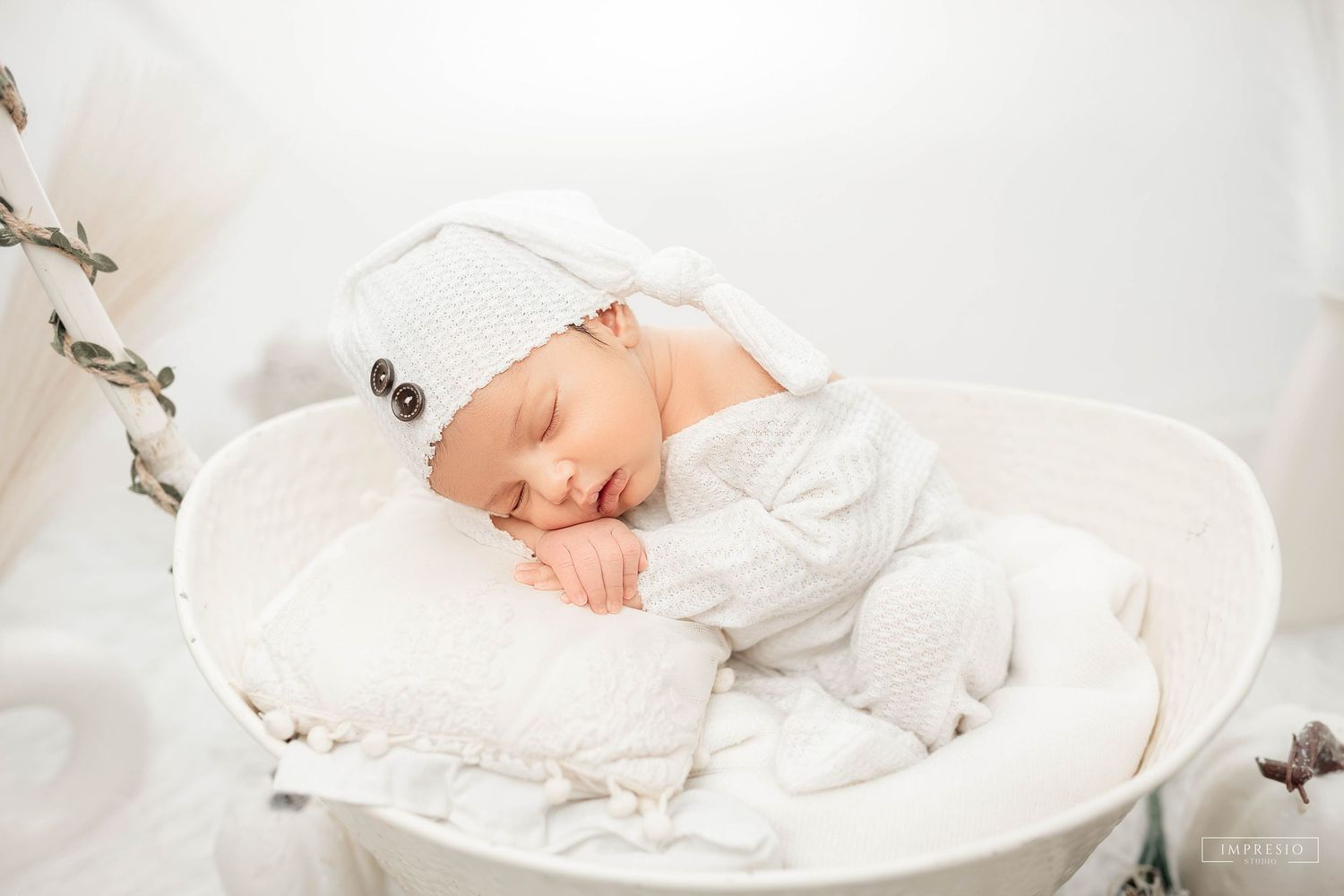 Peaceful newborn baby sleeping in white basket wearing white onesie and knit hat, soft light nursery photography.