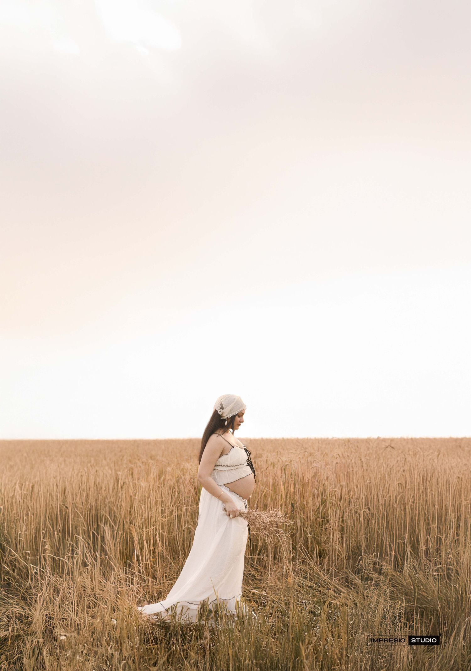 Pregnant woman in white dress holding wheat stalks in golden field at sunset.