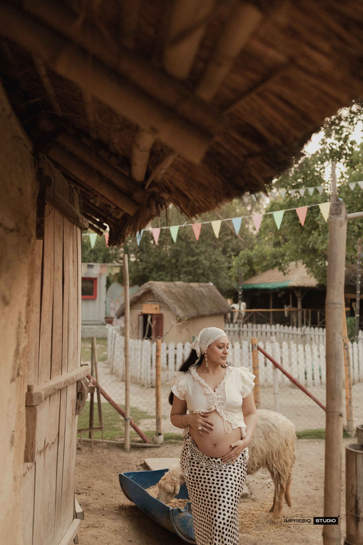 Pregnant woman in white top and polka dot skirt cradling her belly on a rustic farm with colorful bunting flags.