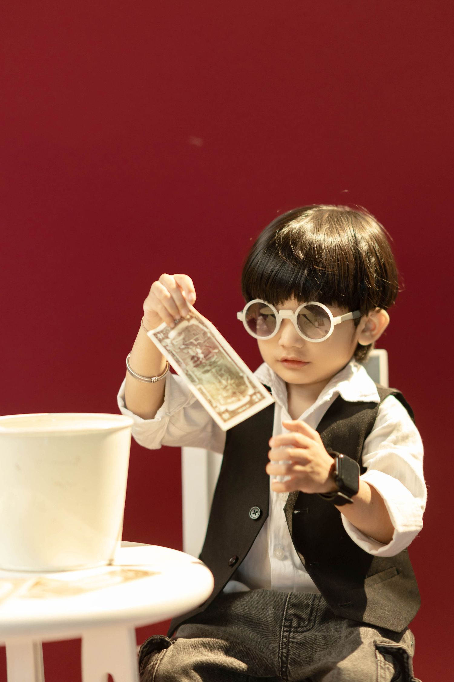 Young child dressed in vest and round glasses holding a dollar bill near a white top hat on a table.