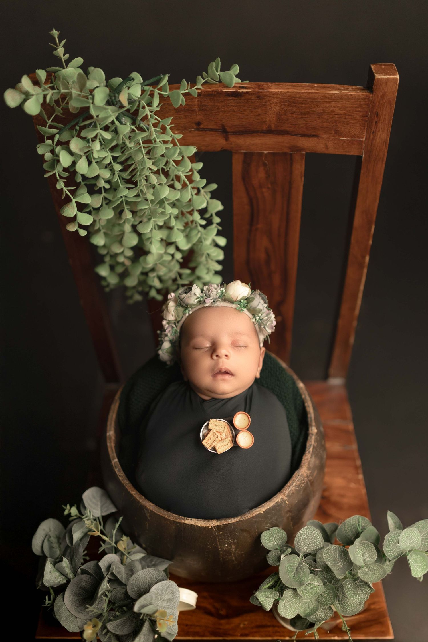 Newborn baby sleeping in a wooden bowl wrapped in dark green fabric, wearing a floral bonnet, surrounded by eucalyptus leaves.