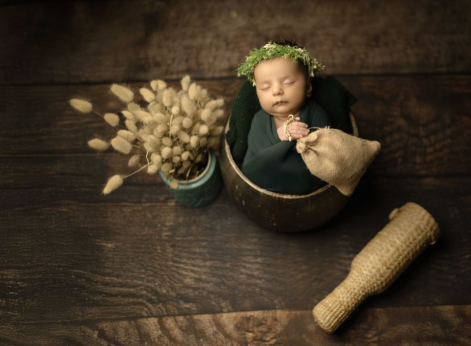 Newborn baby sleeping in a wooden bowl with a green floral crown, wrapped in teal fabric, beside dried flowers on rustic wood.
