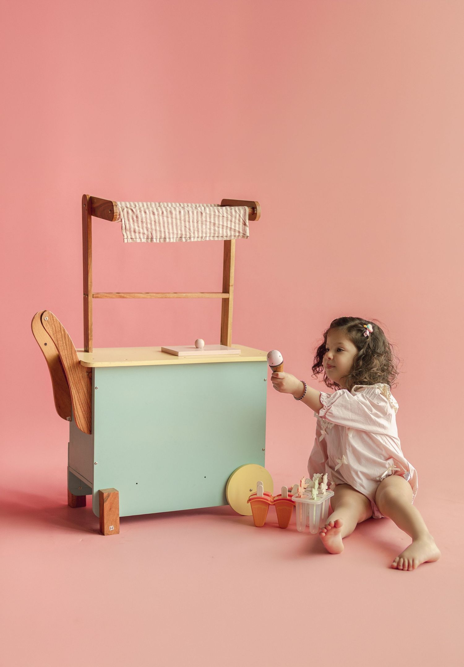 Toddler girl playing with a wooden toy ice cream cart and accessories on a pink background.