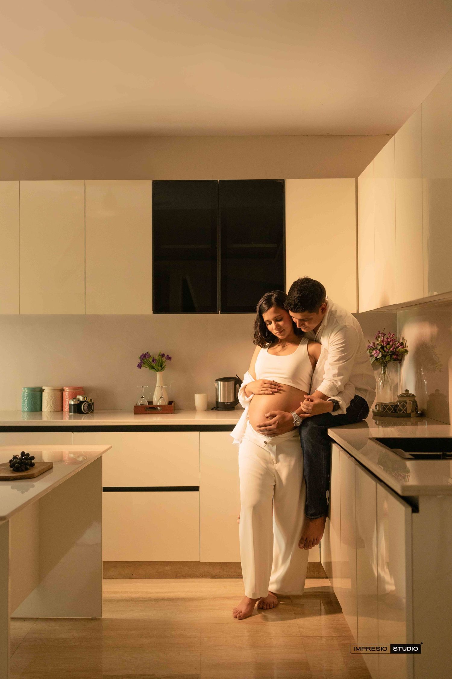 A couple sharing a tender moment in a modern kitchen, with the woman proudly displaying her pregnant belly.