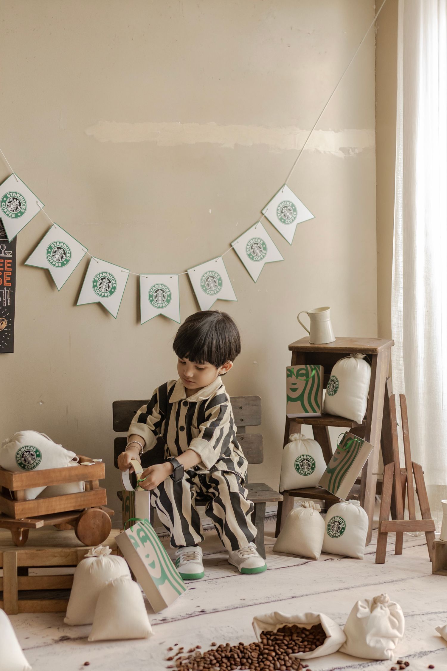 Young boy in striped outfit plays with Starbucks-themed burlap bags among coffee beans in a decorated room.