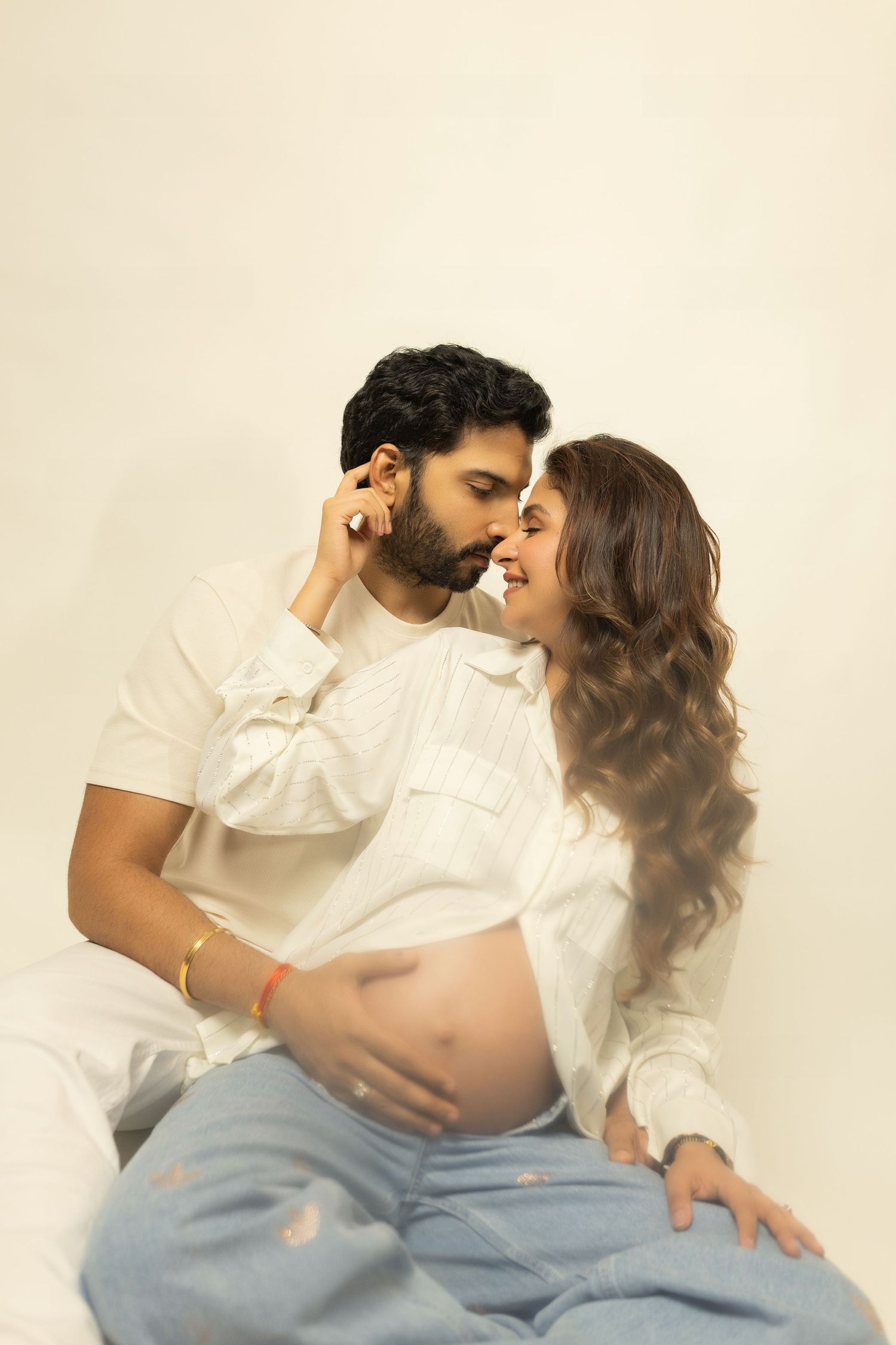 Couple in white outfits sharing a tender moment, pregnant woman cradling her baby bump in a maternity photoshoot.