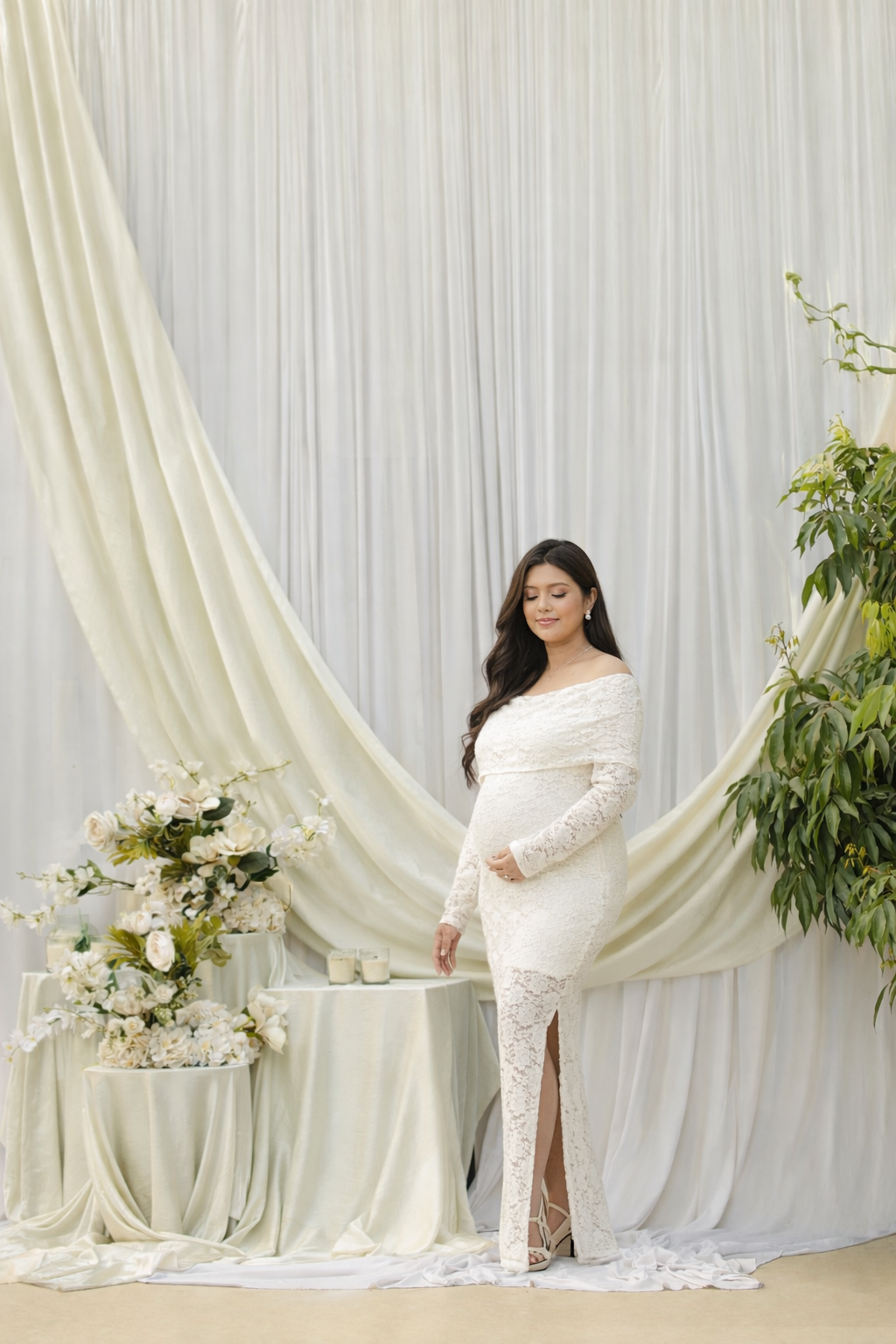 A maternity photoshoot featuring elegant white drapes, floral arrangements, and a flowing white lace gown.