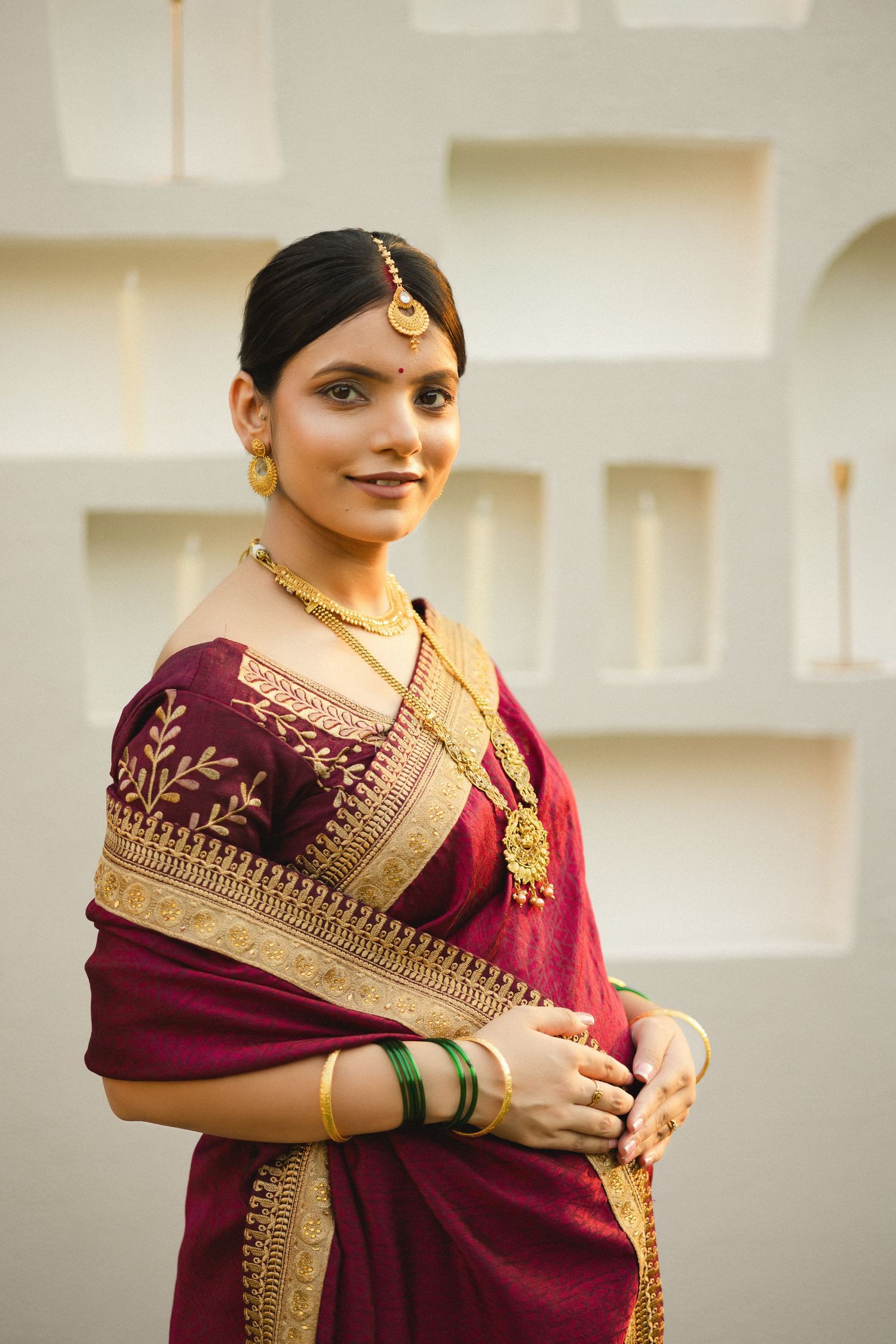 Woman wearing traditional burgundy saree with ornate gold border and jewelry poses gracefully in soft natural light.