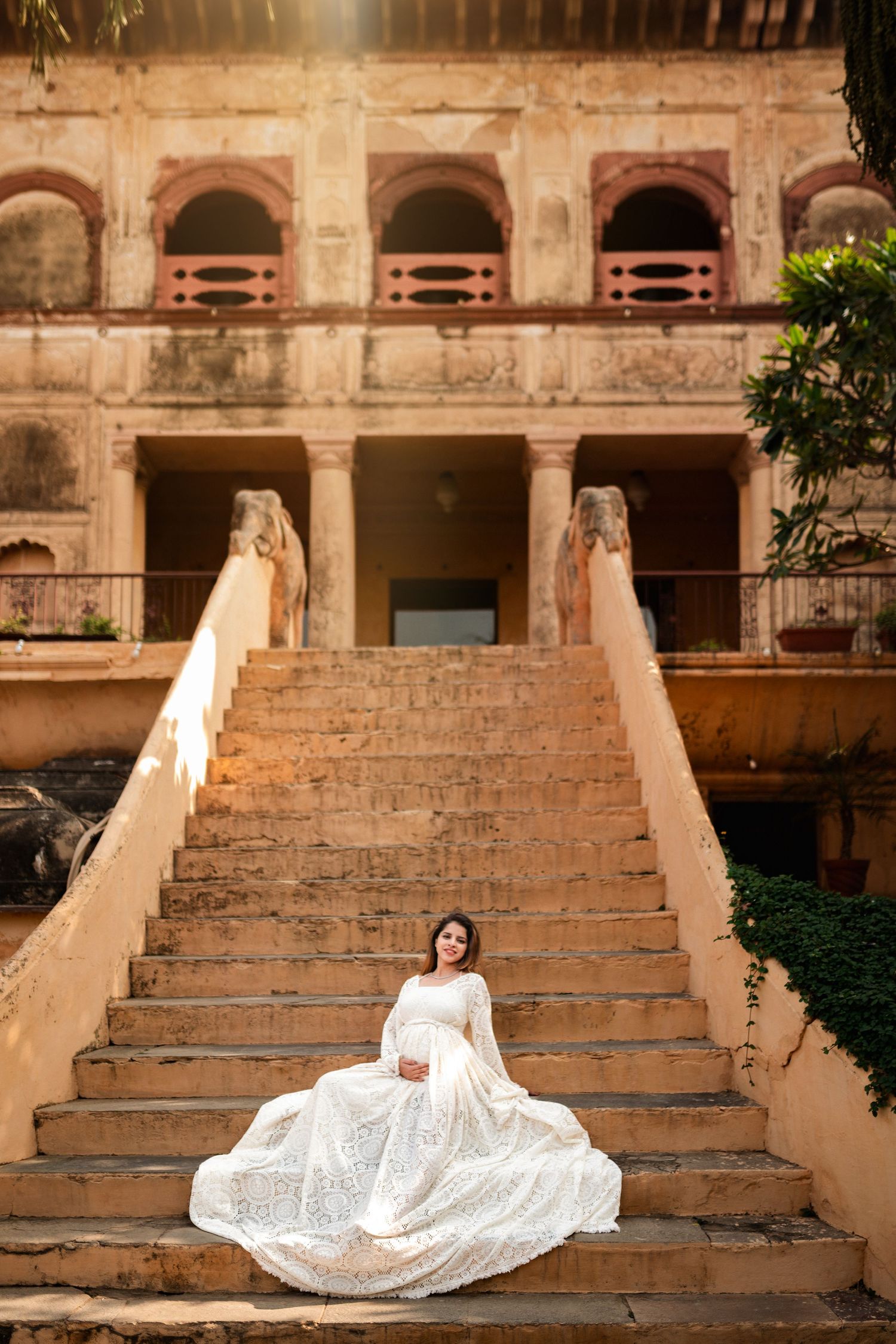 Pregnant woman in white lace gown sitting on stone steps of an ornate historic building in warm sunlight.
