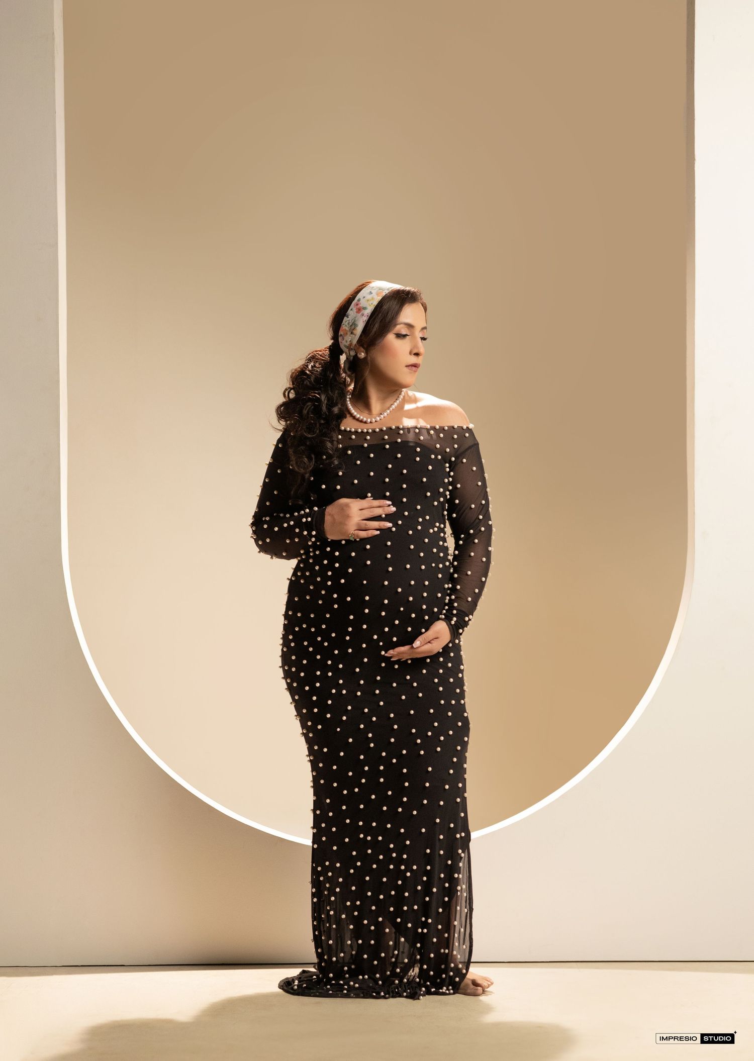 Pregnant woman in black polka dot off-shoulder gown poses elegantly before an arched backdrop.