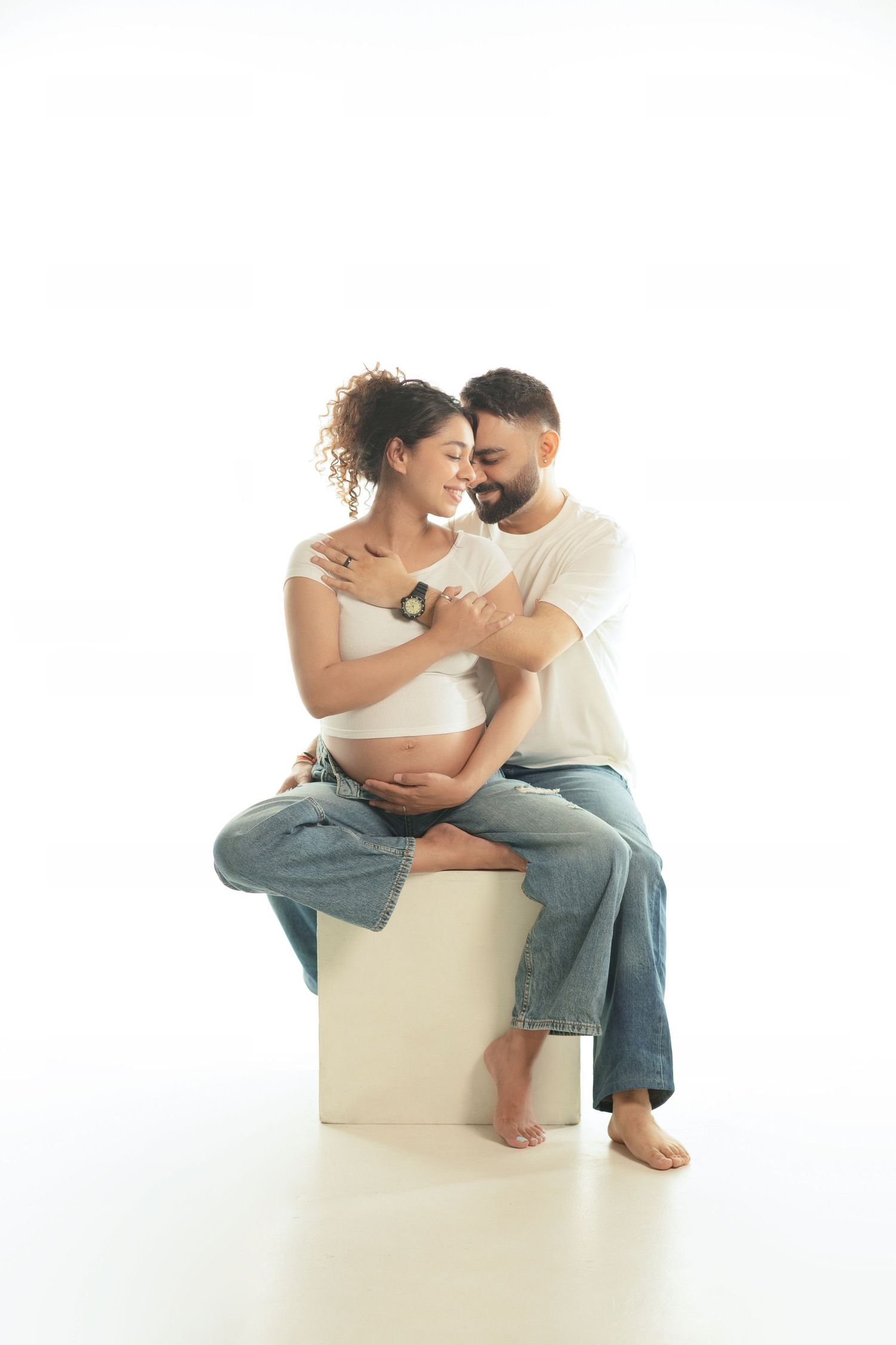 Happy couple embracing tenderly, pregnant woman sitting on white cube with partner behind her, both wearing jeans.