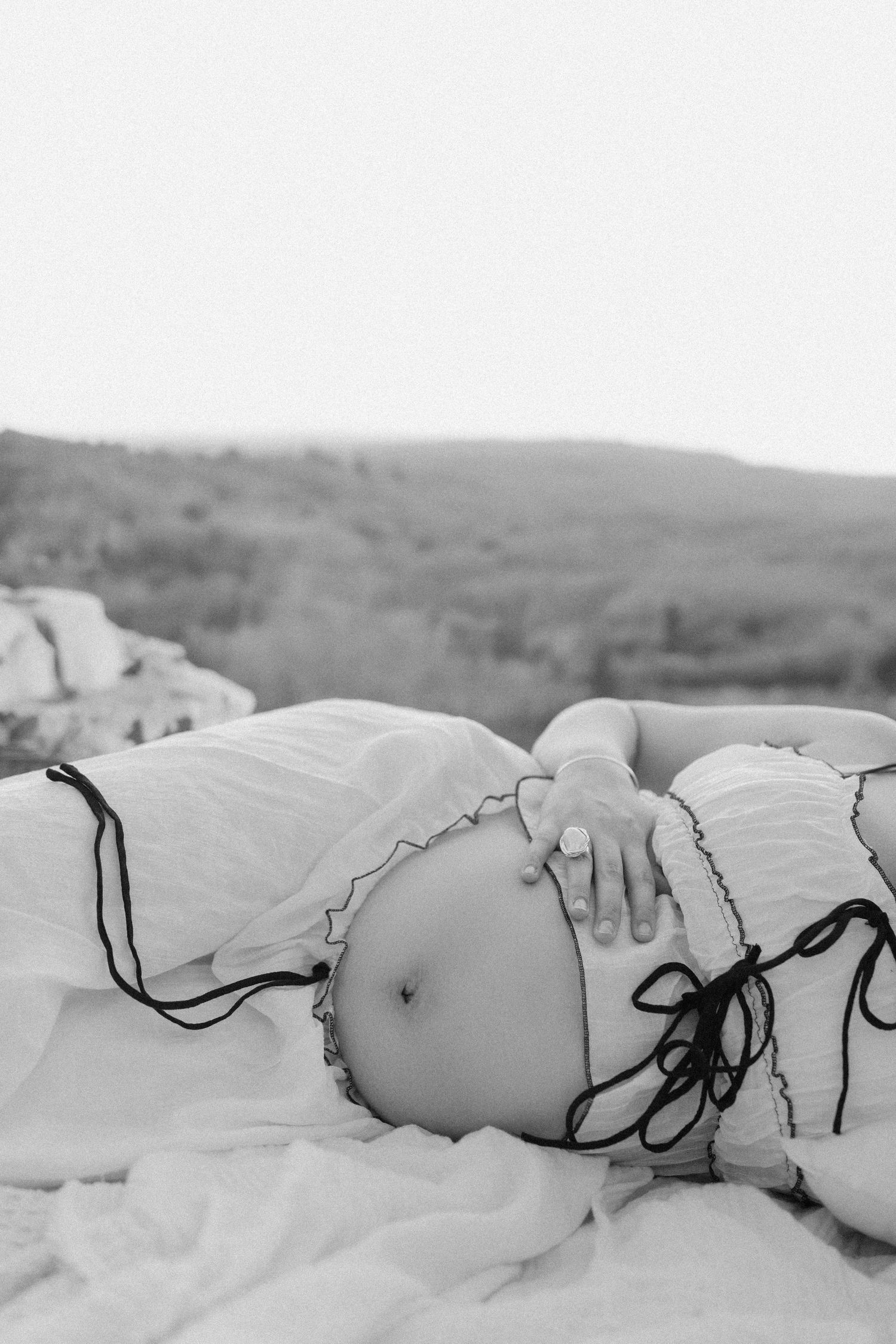Black and white maternity photo of pregnant woman lying outdoors, showing bare belly with hand resting on bump.