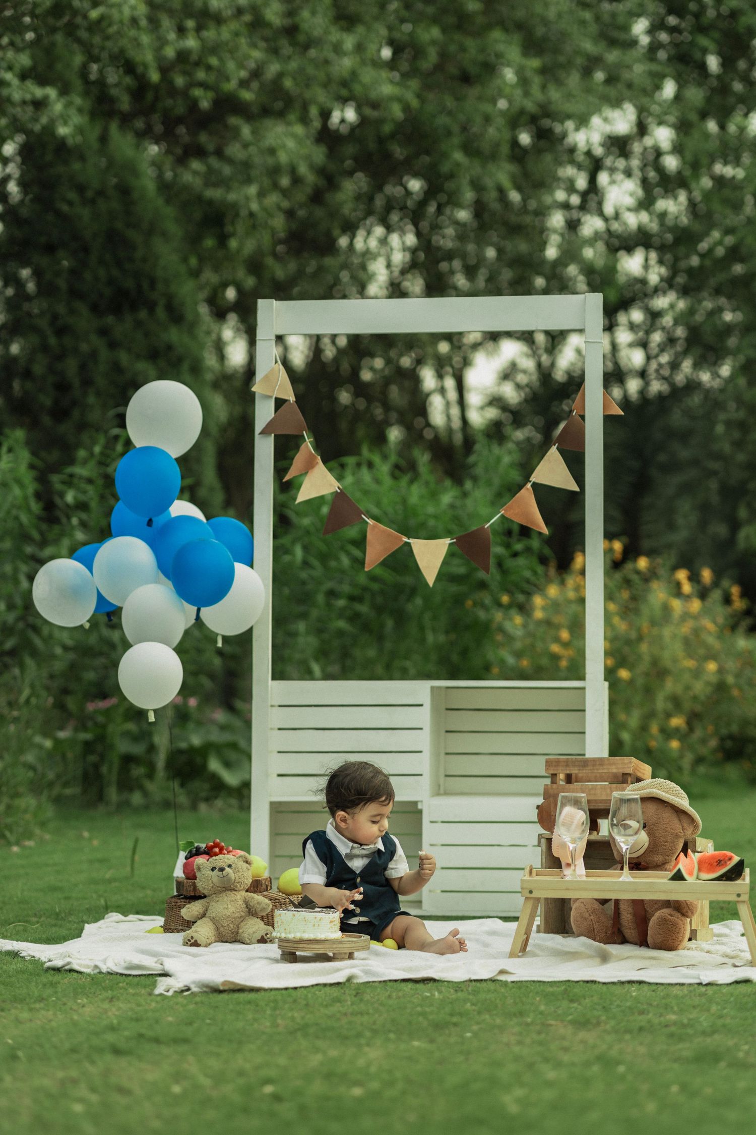 Baby boy sitting outdoors at a first birthday picnic setup with balloons, teddy bears, and bunting decorations.