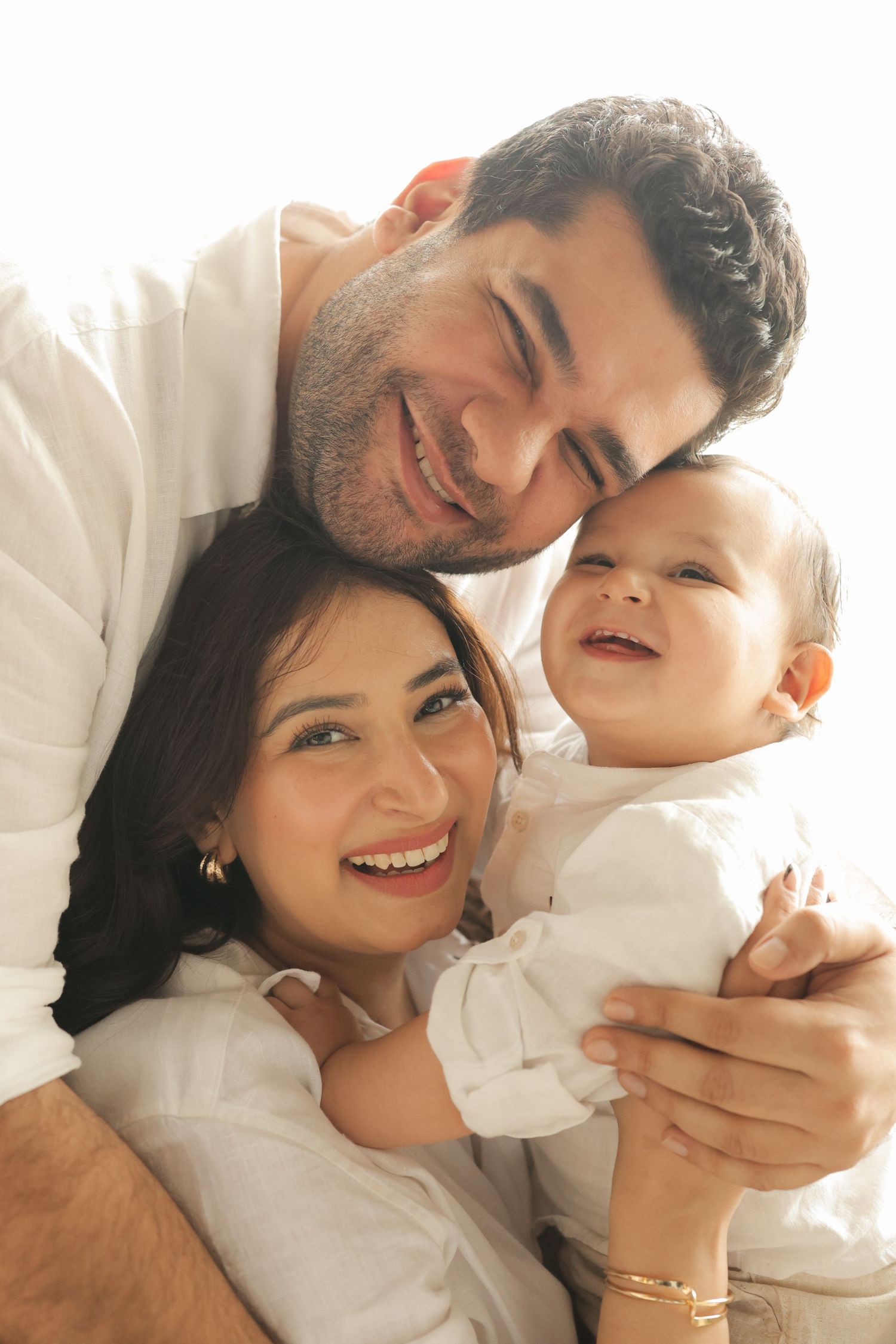 Happy family portrait with smiling parents holding their joyful baby, all dressed in white.