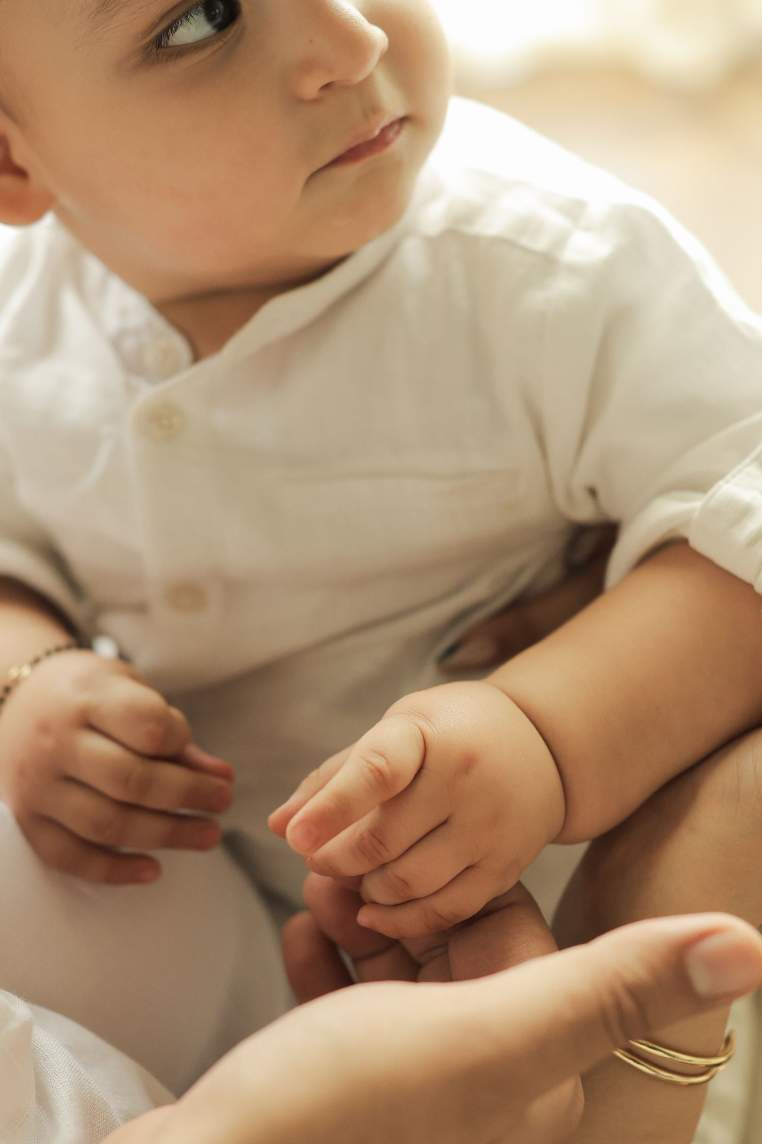 A baby in a white outfit holds an adult's hand, wearing a gold bracelet, in a soft, warm-toned setting.