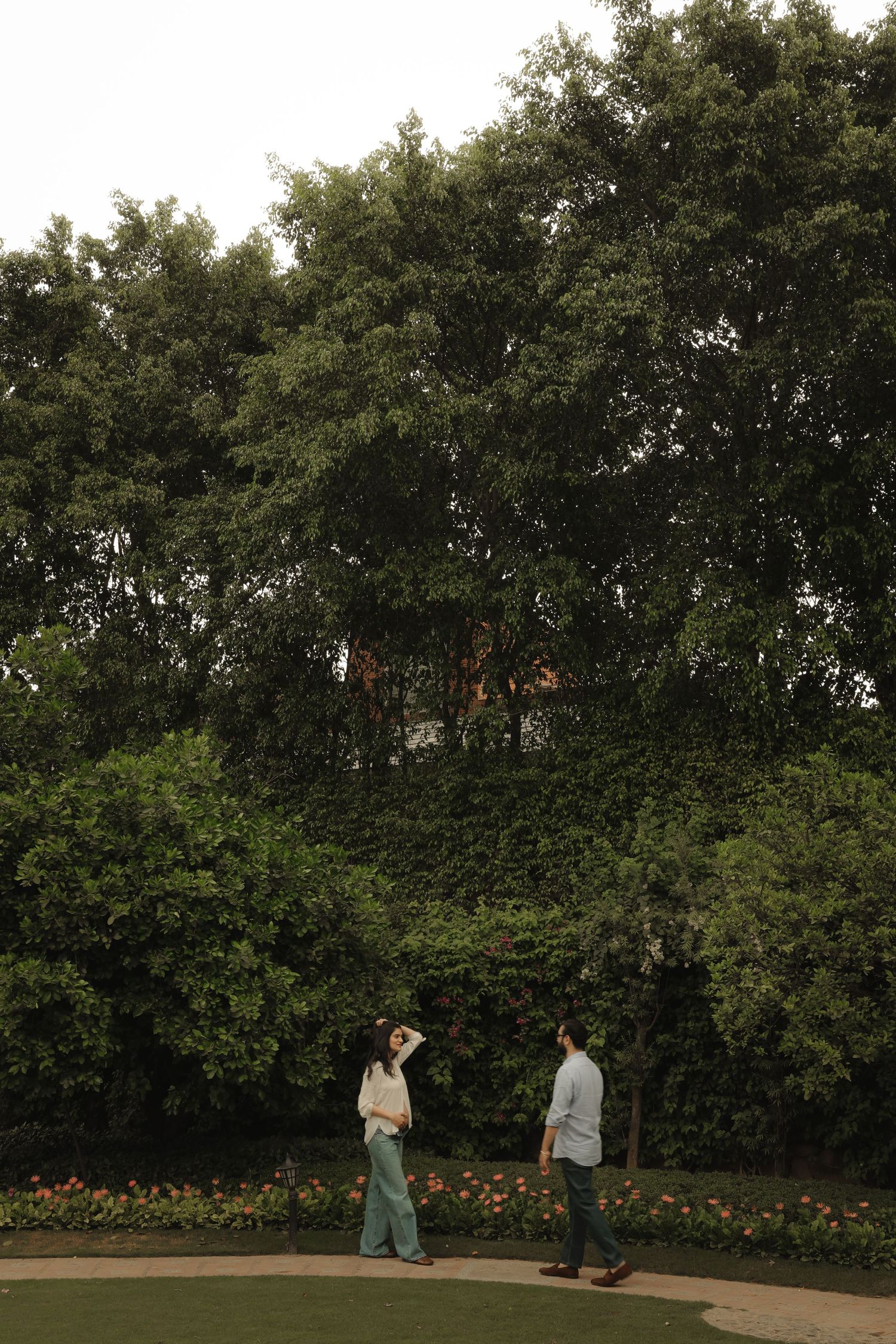 A couple stands facing each other in a lush green garden park surrounded by tall trees and colorful flowers.