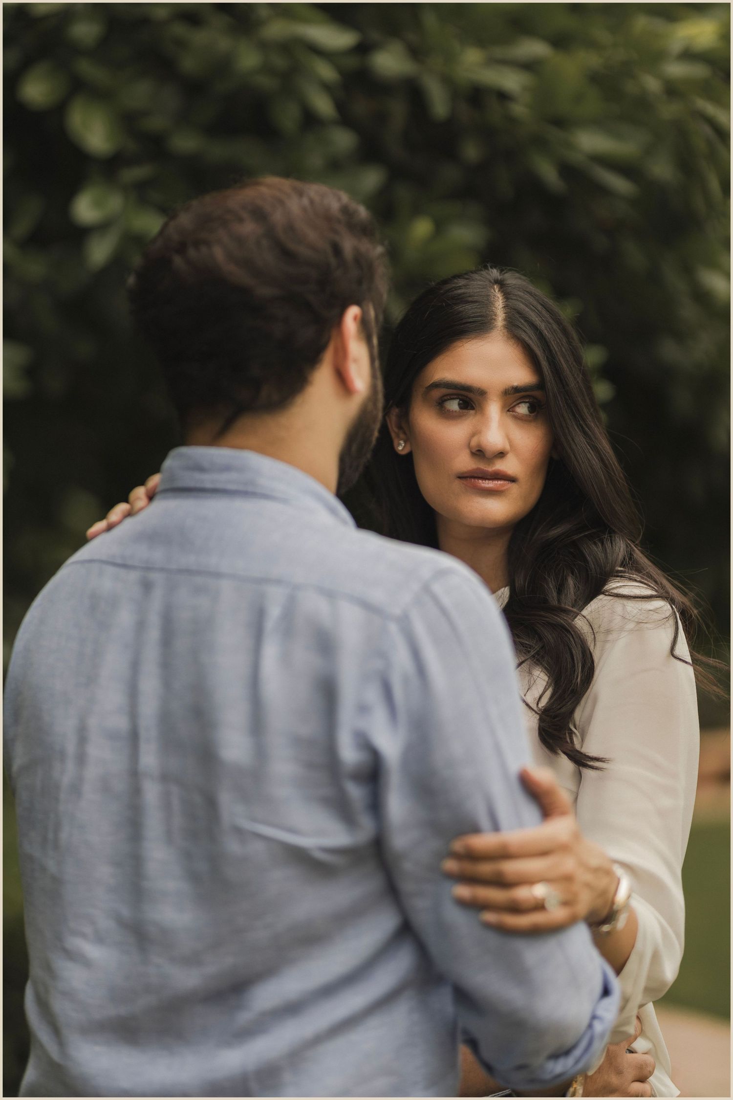 Couple sharing an intimate moment outdoors, woman in white top gazing at man in blue shirt amid lush greenery.