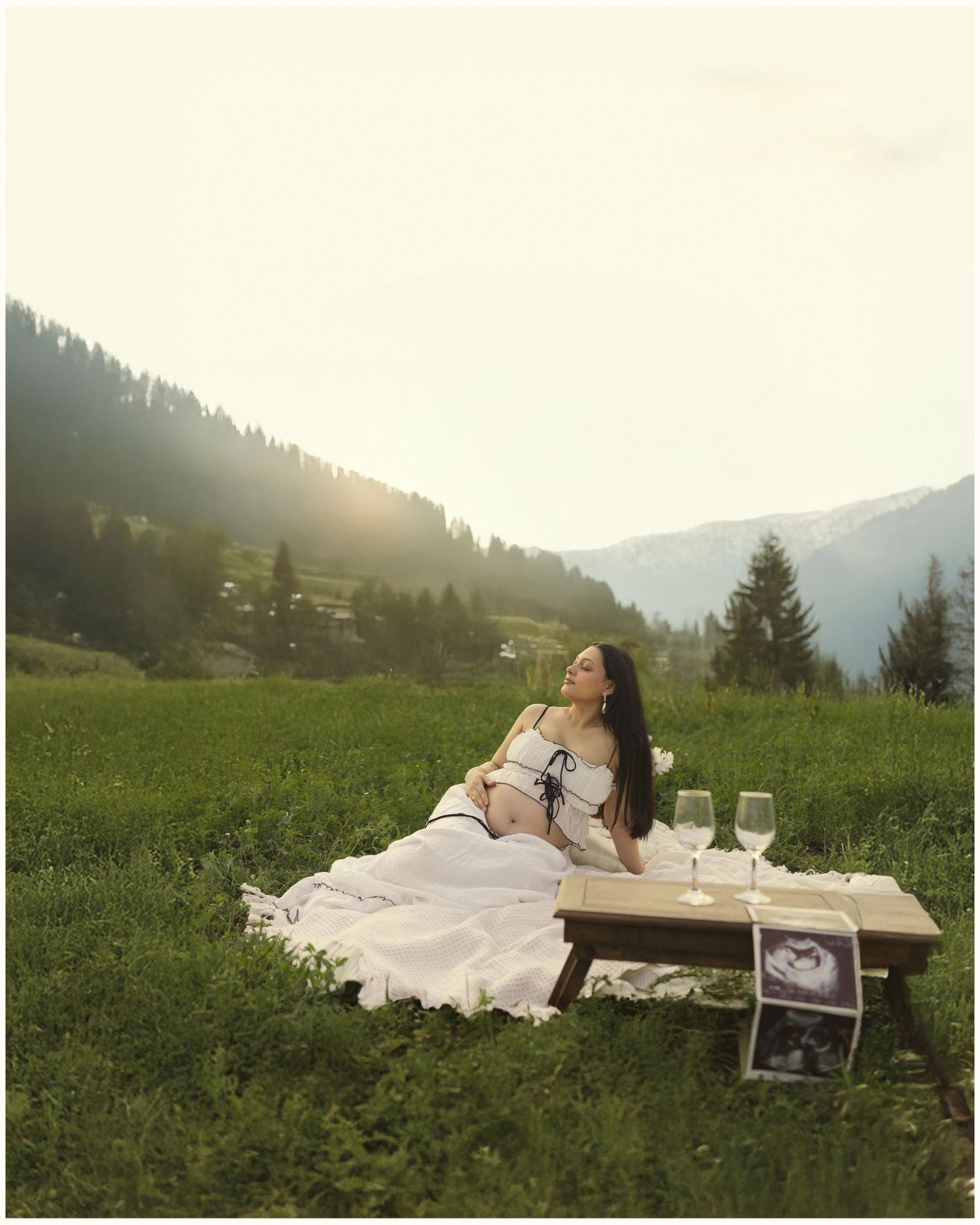 Pregnant woman in white outfit reclines on wooden bench in alpine meadow at sunset with ultrasound photos and wine glasses.