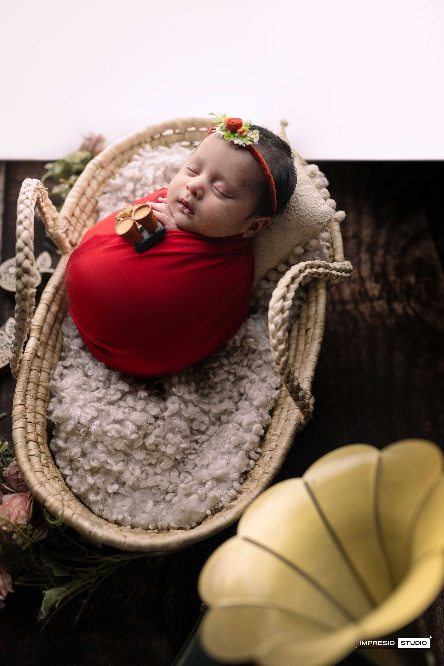 Newborn baby swaddled in red wrap, sleeping in a wicker basket with floral headband, professional studio photo.