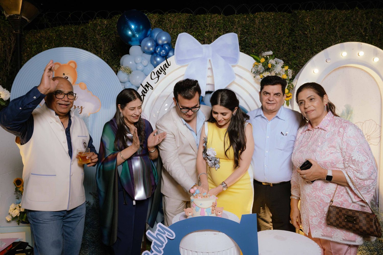 Group of six people cutting a cake at a baby shower celebration with blue balloon decorations.
