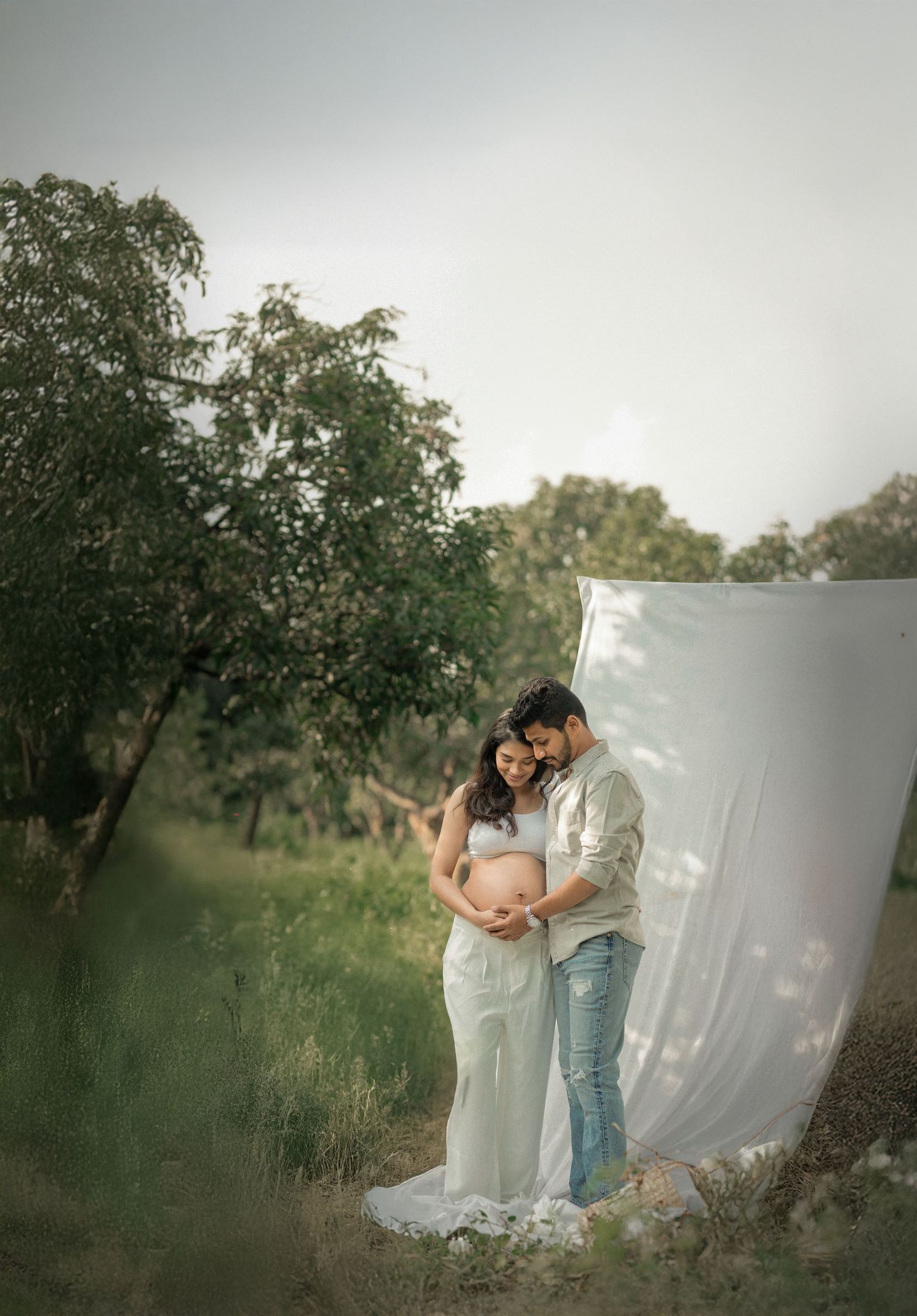A couple shares an intimate moment during their maternity photoshoot outdoors with a white backdrop and natural scenery.